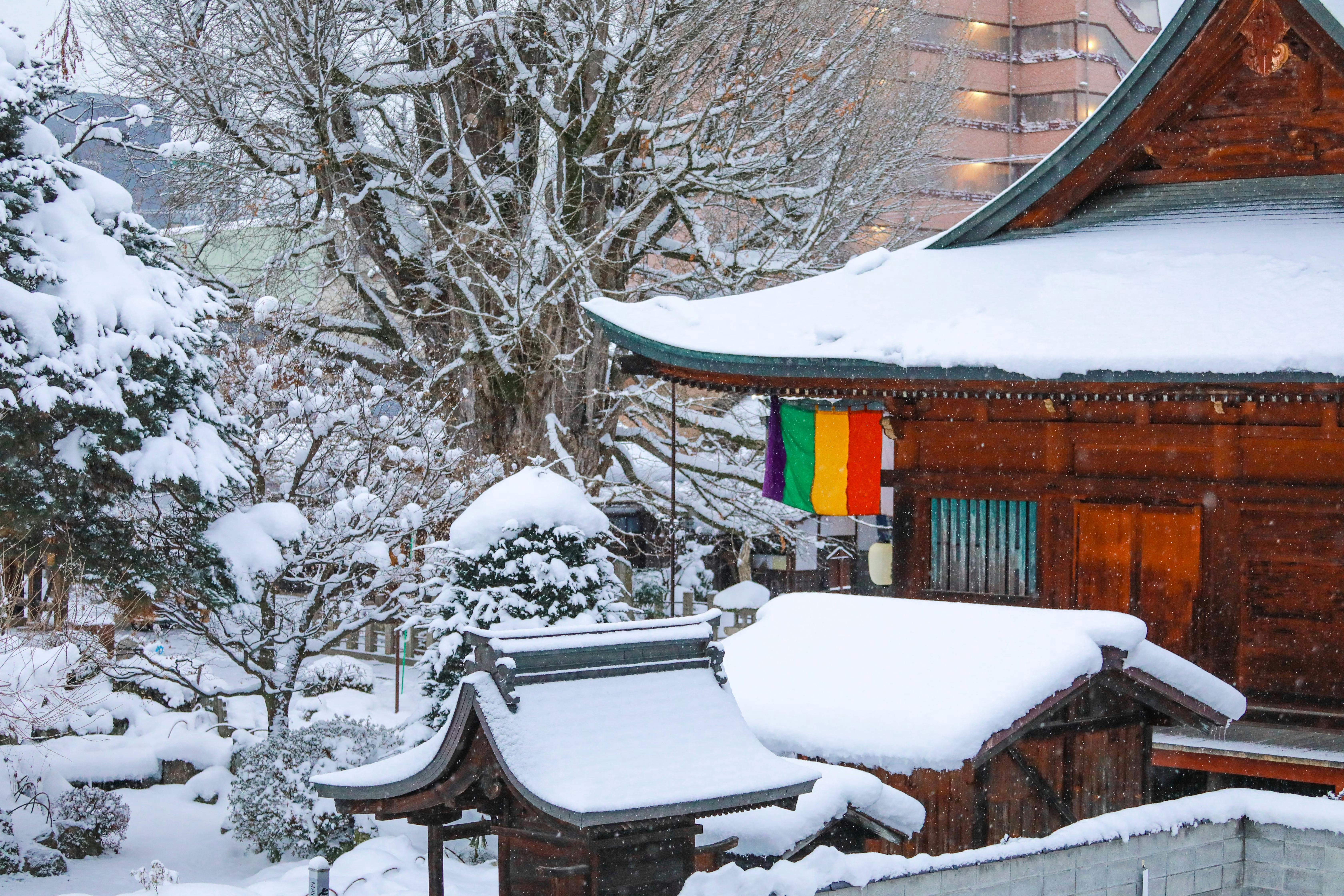 A traditional wooden Japanese building and garden covered in heavy snow, with a colorful five-striped flag hanging from the roof and bare trees in the background.