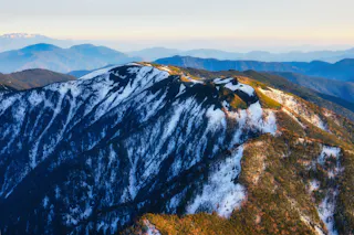 A scenic mountain range with snow-covered peaks and forested slopes, bathed in warm sunlight. Layers of distant blue mountains fade into the horizon under a clear sky.