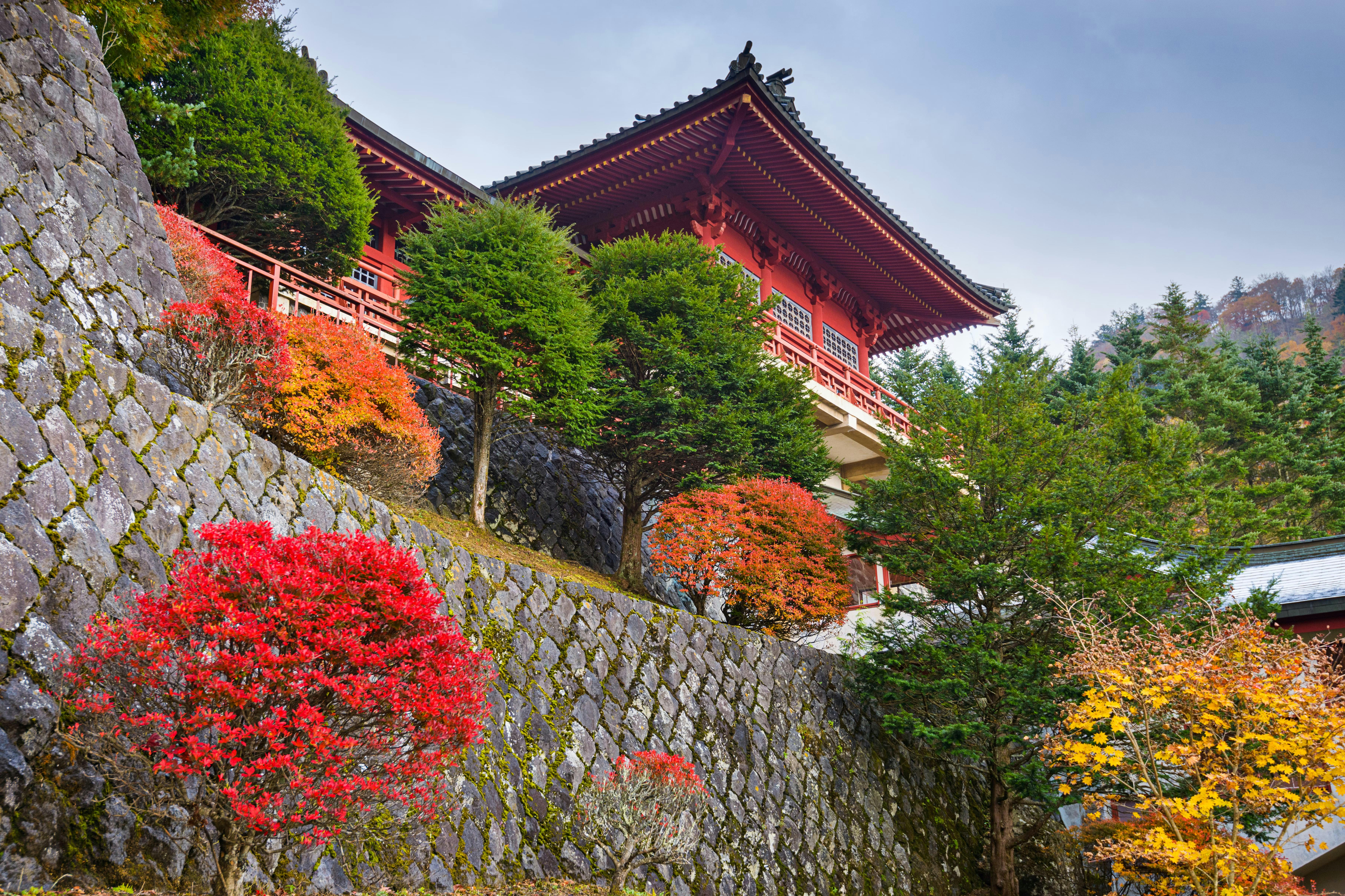 A traditional Japanese building with red wooden accents sits on a stone terrace, surrounded by lush trees with vibrant autumn foliage in red, orange, and yellow hues under a cloudy sky.