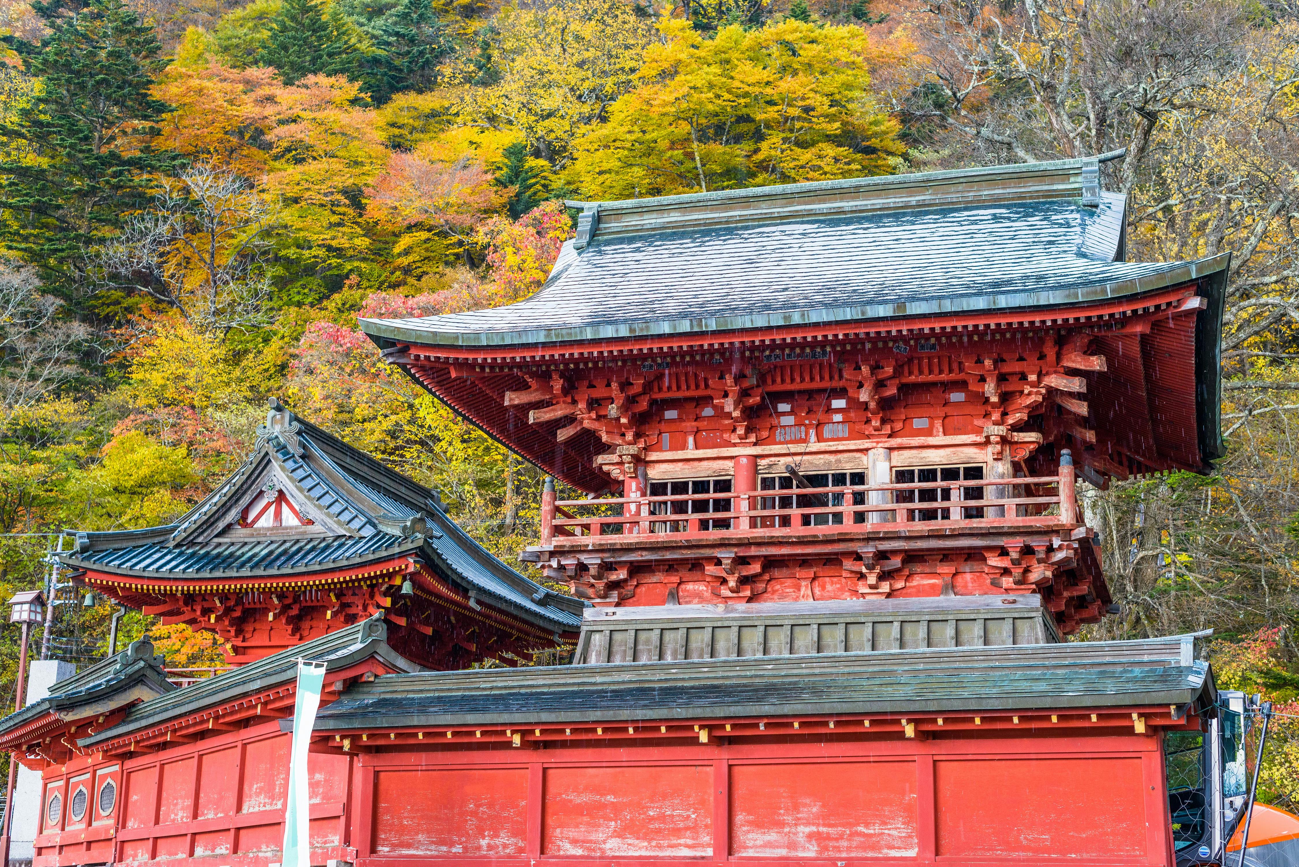 A traditional red Japanese temple with ornate wooden architecture surrounded by colorful autumn trees with yellow, orange, and green leaves.