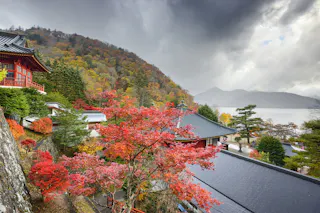 A scenic view of a traditional Japanese temple surrounded by vibrant red autumn leaves, with forested hills and a lake in the background under a dramatic cloudy sky.