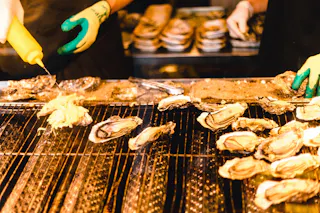 Two people wearing gloves prepare oysters on a grill; one person squeezes sauce from a yellow bottle onto the oysters, with many shells cooking on the hot surface.