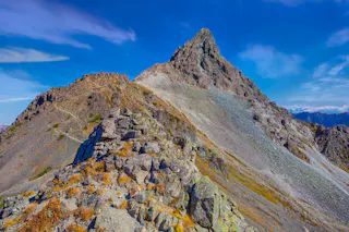Rocky mountain peak with jagged ridges under a bright blue sky, with patches of dry grass and loose stones along the slopes. A rugged trail winds up toward the summit amidst dramatic alpine scenery.