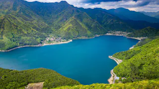 A deep blue lake is surrounded by lush green mountains under a partly cloudy sky, with winding roads and small buildings along the shoreline.