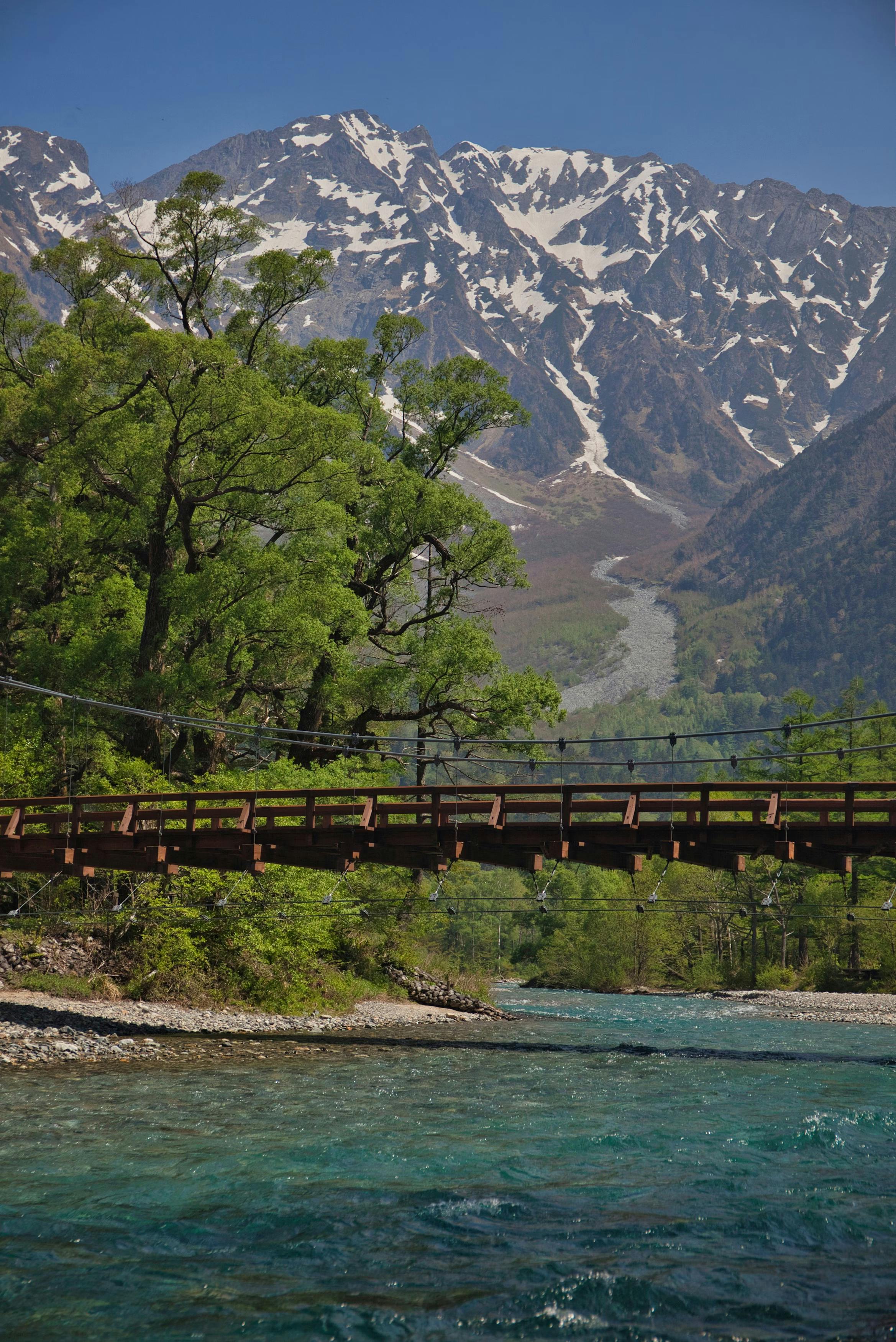 A wooden bridge crosses a clear blue river with lush green trees on the banks. In the background, snow-capped mountains rise under a blue sky.