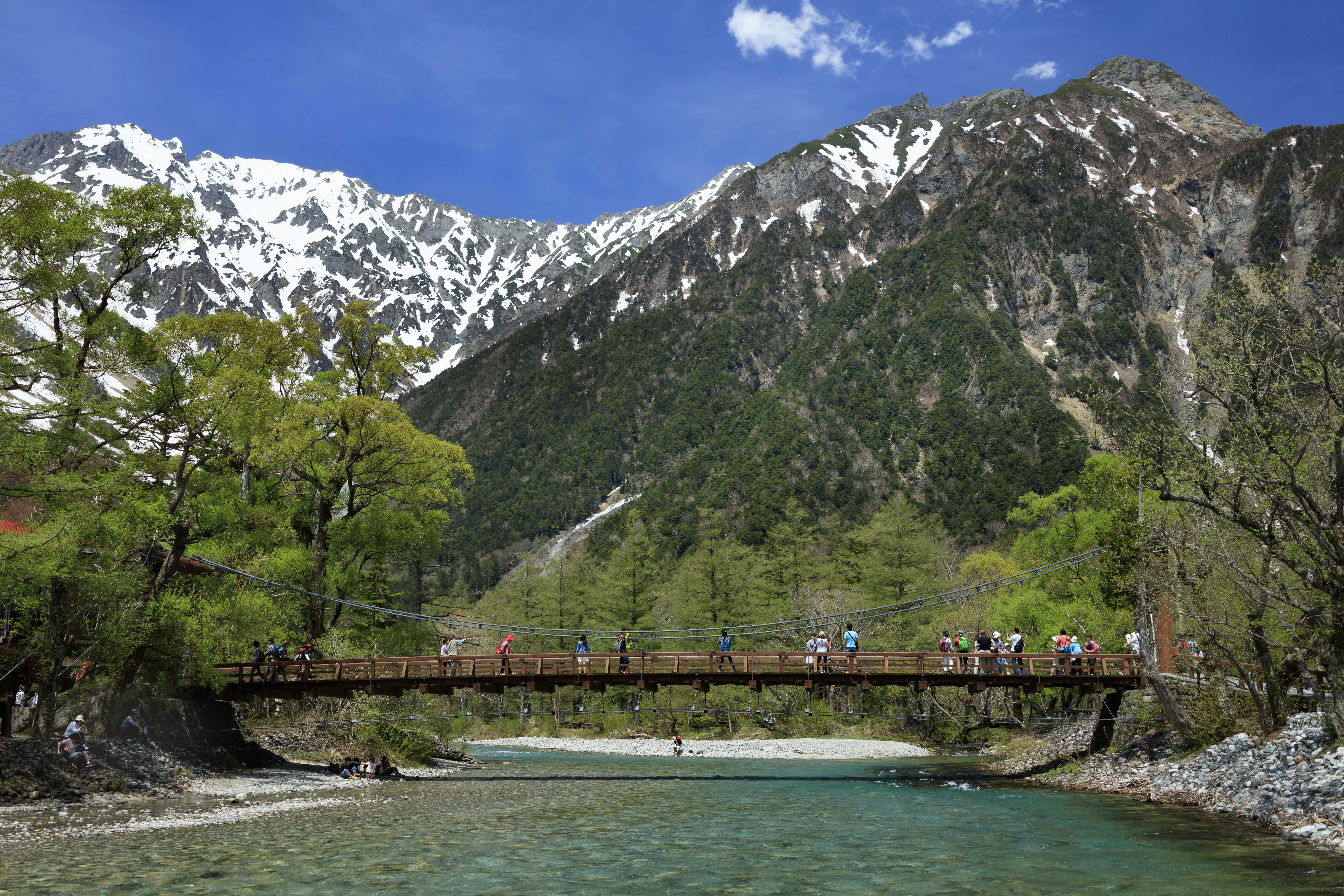 A wooden suspension bridge crosses a clear river, with people walking on it. Behind the bridge are tall green trees and snow-capped mountains under a bright blue sky.