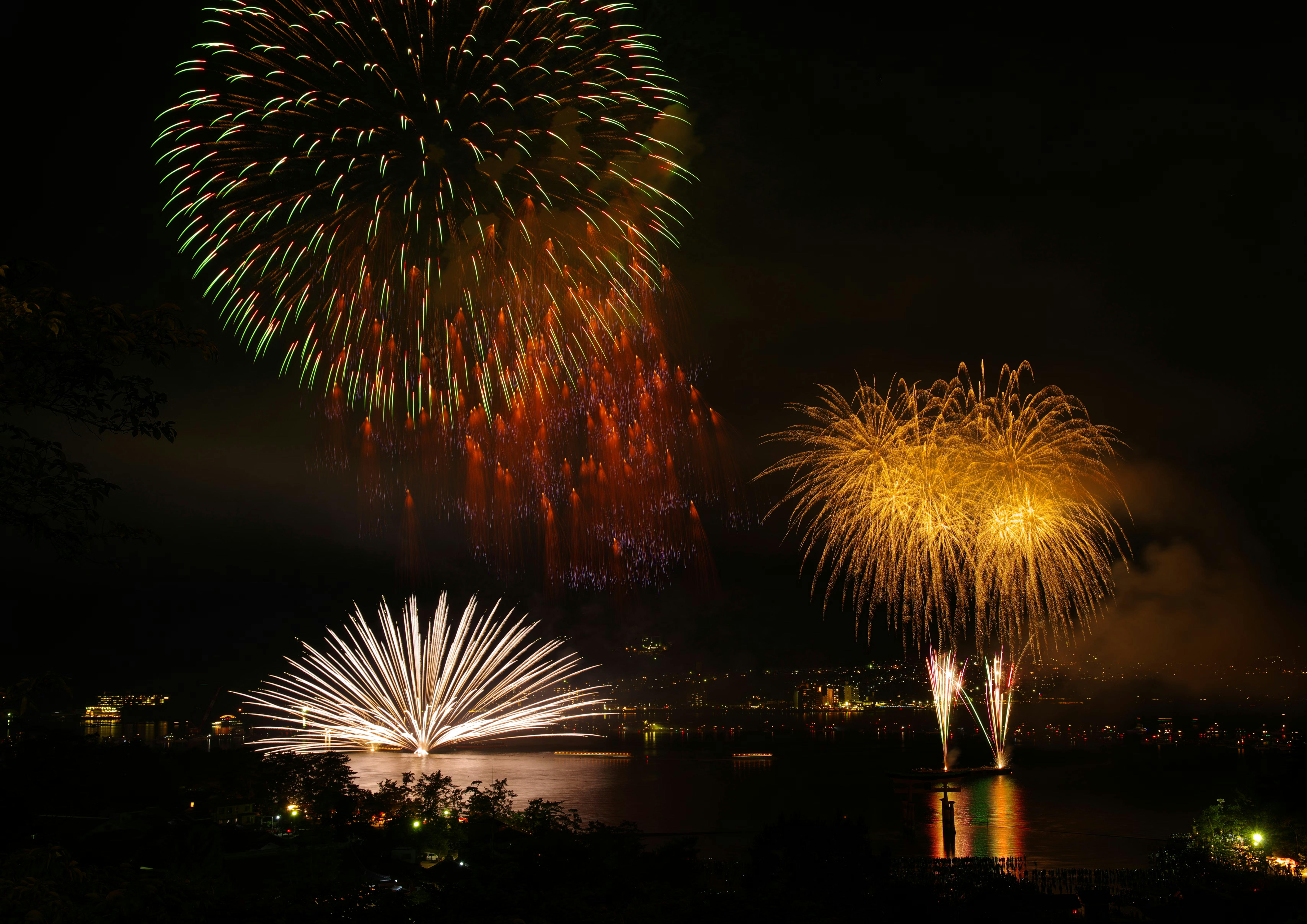 Colorful fireworks burst and light up the night sky over a body of water, reflecting on the surface below. The silhouette of trees and city lights are visible in the background.