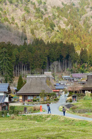 A rural Japanese village with traditional thatched-roof houses surrounded by lush green trees and hills. A few people walk along a winding road through the village.
