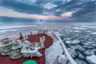View from the bow of a ship navigating through sea ice under a cloudy sky at sunset or sunrise, with a flag flying at the front and scattered ice floes in the water ahead.
