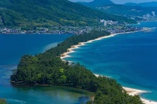A narrow sandbar covered with green trees stretches across blue water, connecting two land areas with a town and mountains in the background under a clear sky.