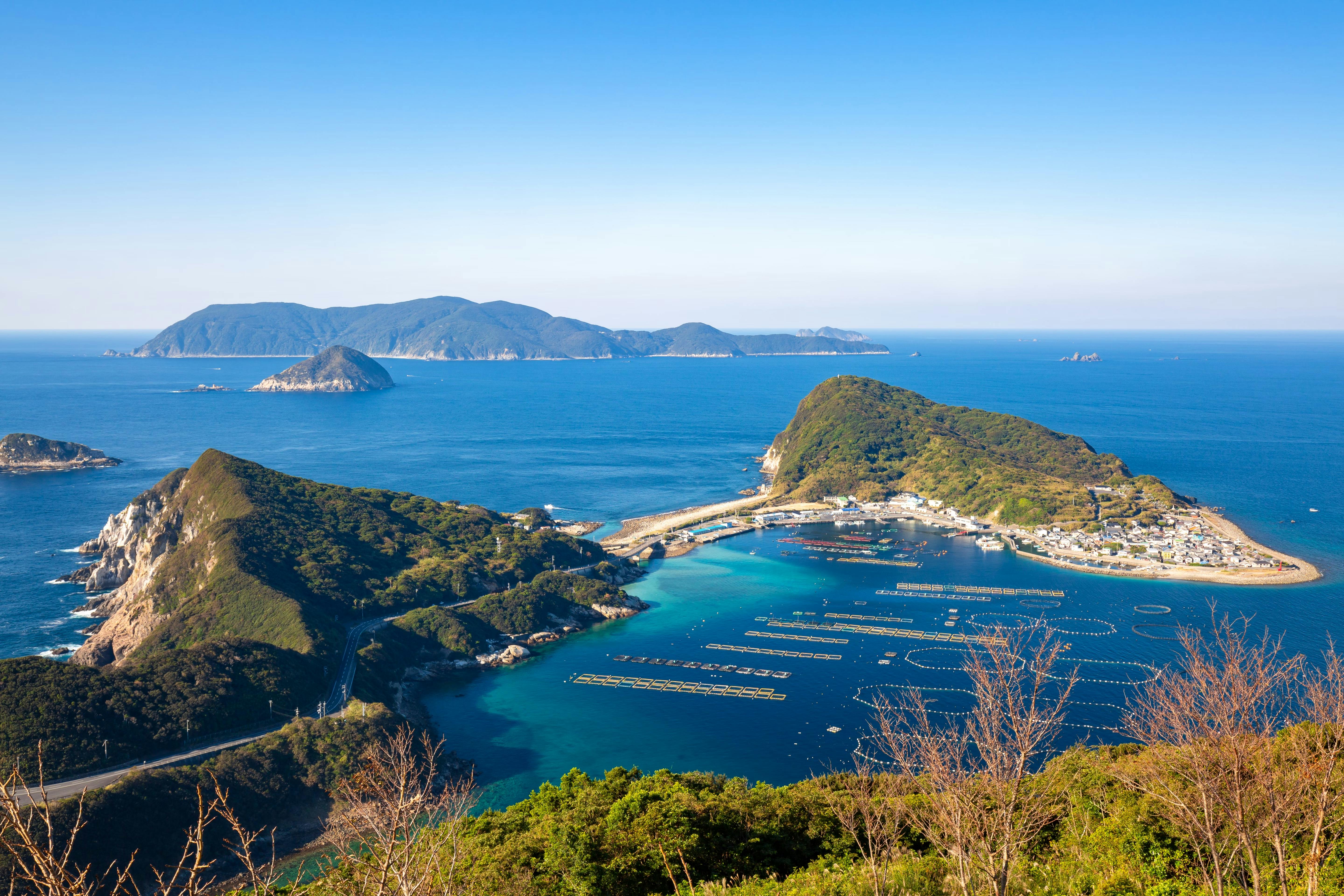 A coastal fishing village sits on a peninsula surrounded by blue sea, with fish farms in the water and green hills in the background. Several islands are visible under a clear, sunny sky.
