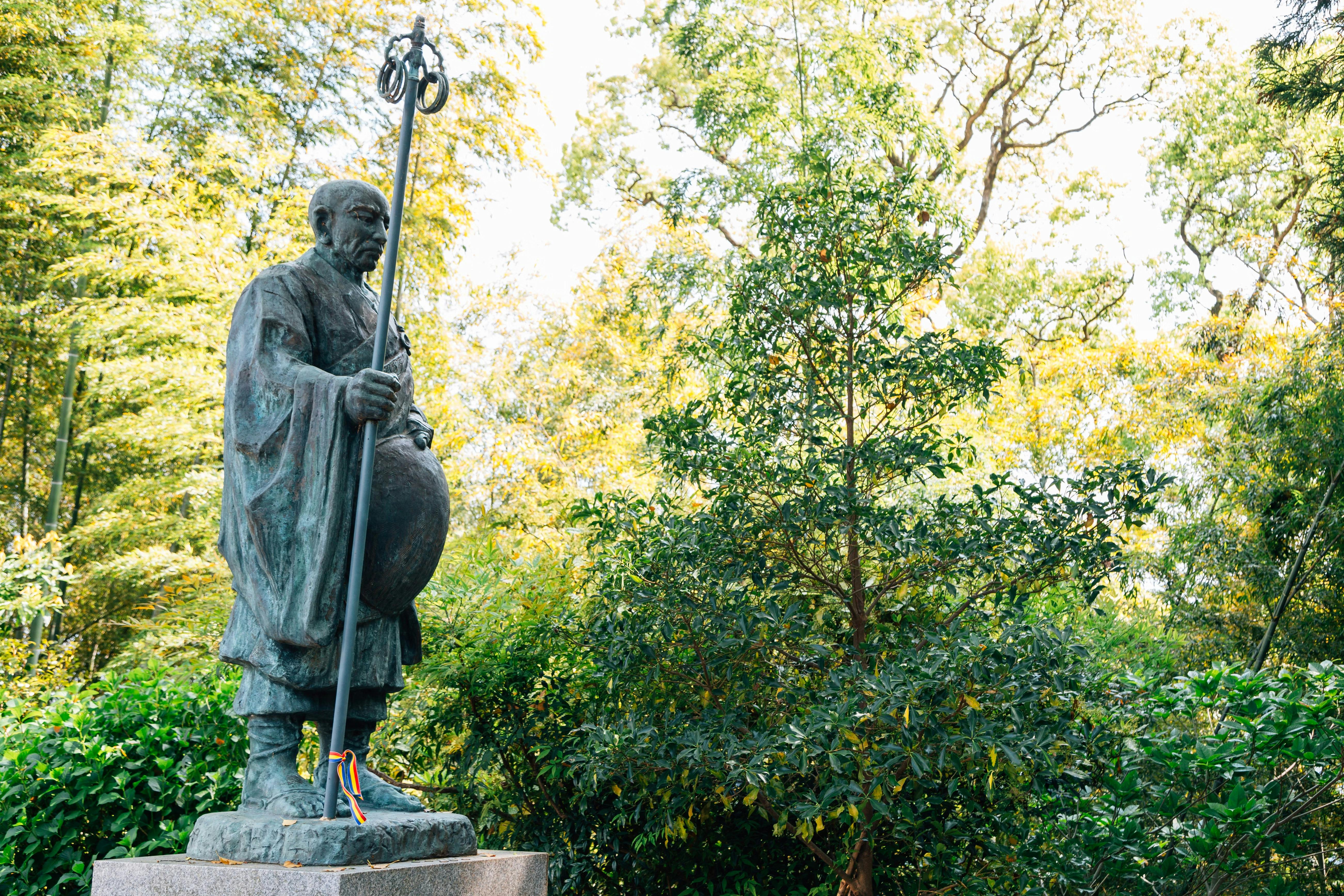 A statue of a robed figure holding a staff stands on a stone pedestal surrounded by lush green trees and foliage in an outdoor garden setting.