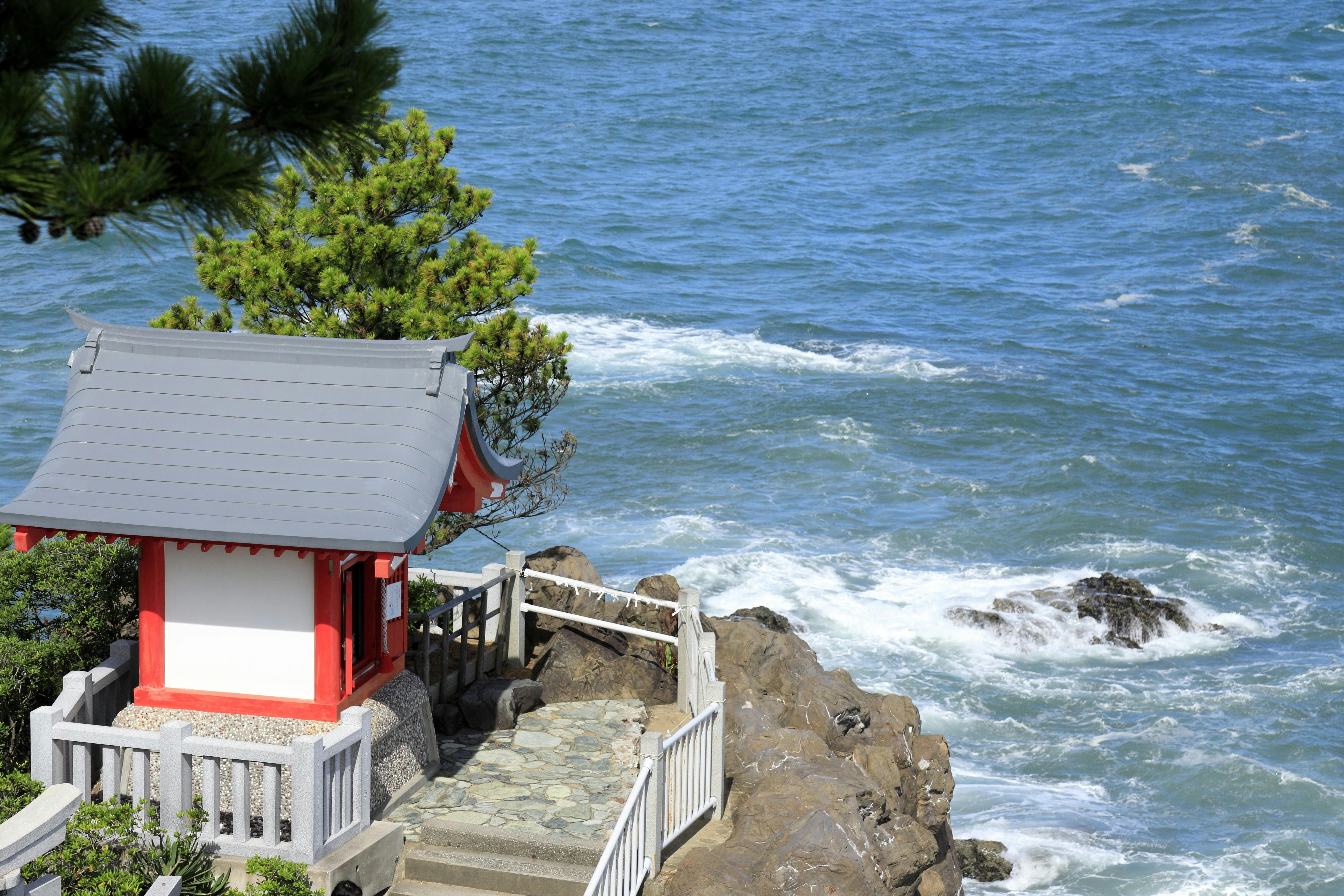 A small red and white shrine sits on a rocky coastline overlooking the blue ocean, with waves crashing against the rocks and pine branches visible in the foreground.