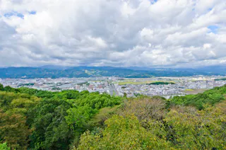 View of a cityscape bordered by lush green trees in the foreground, with mountains in the background under a partly cloudy sky.