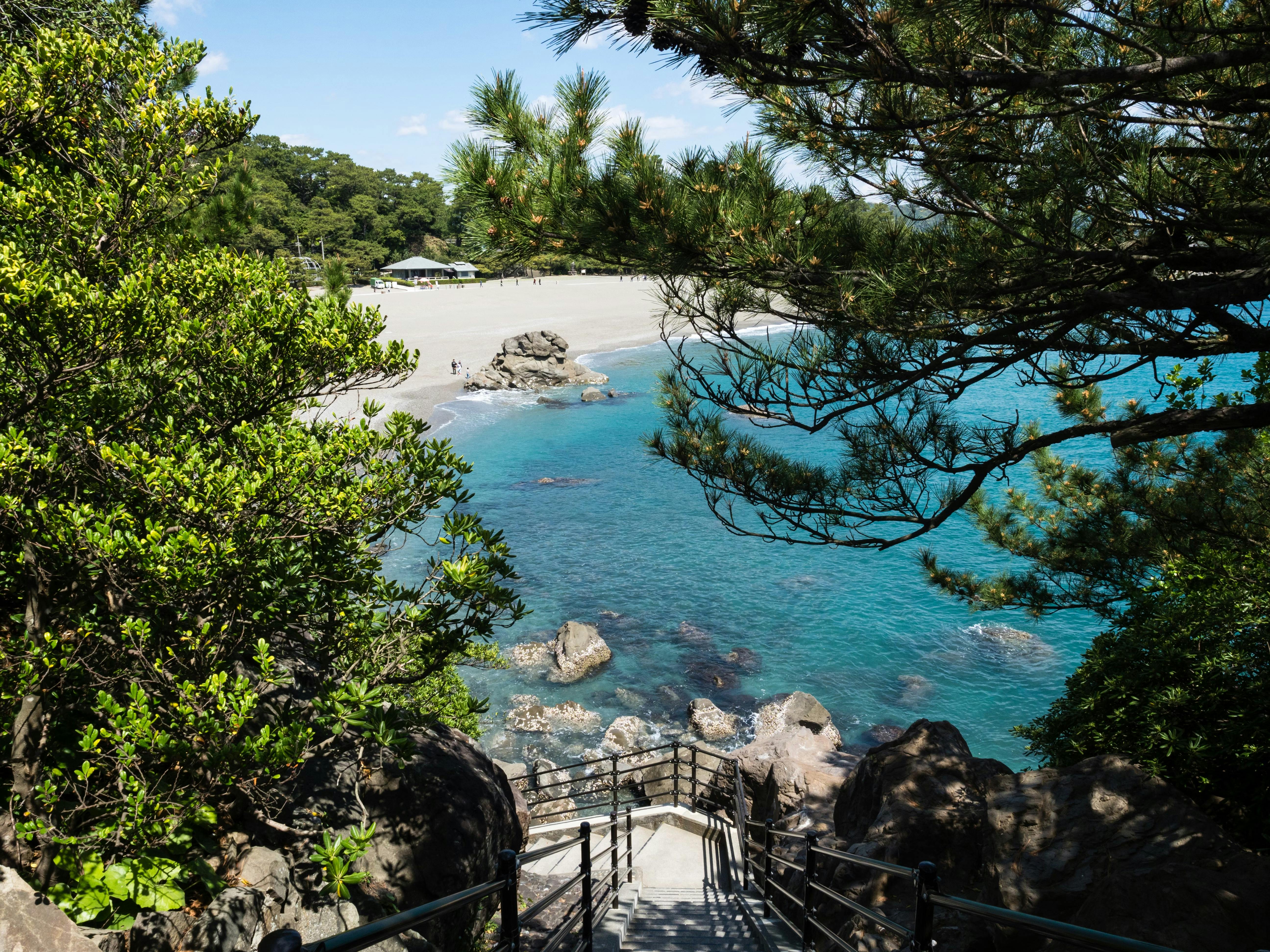 A staircase leads down to a sandy beach with clear blue water, surrounded by green trees and rocks. The sky is mostly clear with a few clouds, and a small shelter is visible in the distance on the beach.