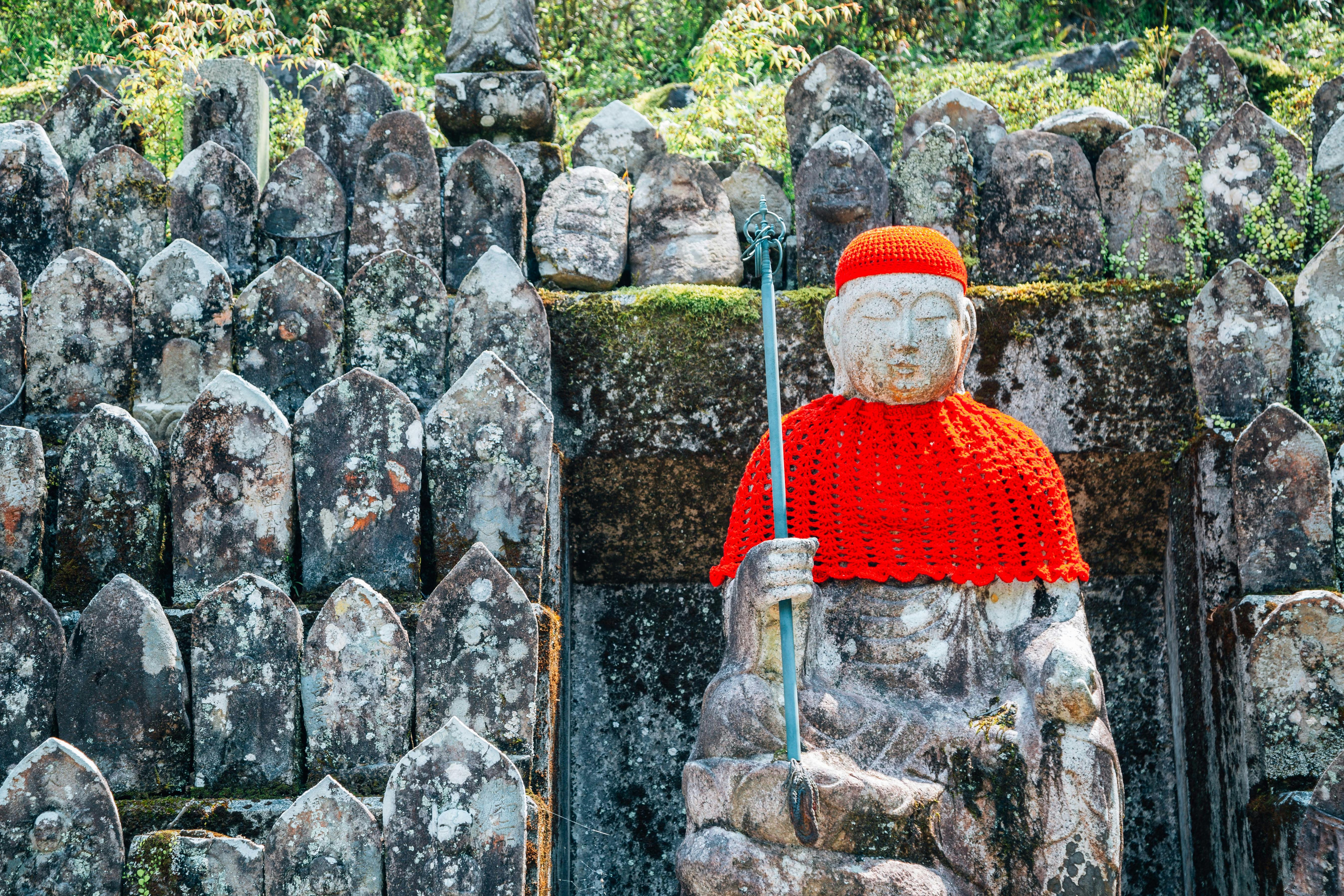 A stone statue dressed in a red crocheted hat and cloak, holding a staff, sits among weathered stone markers, with greenery and moss in the background.