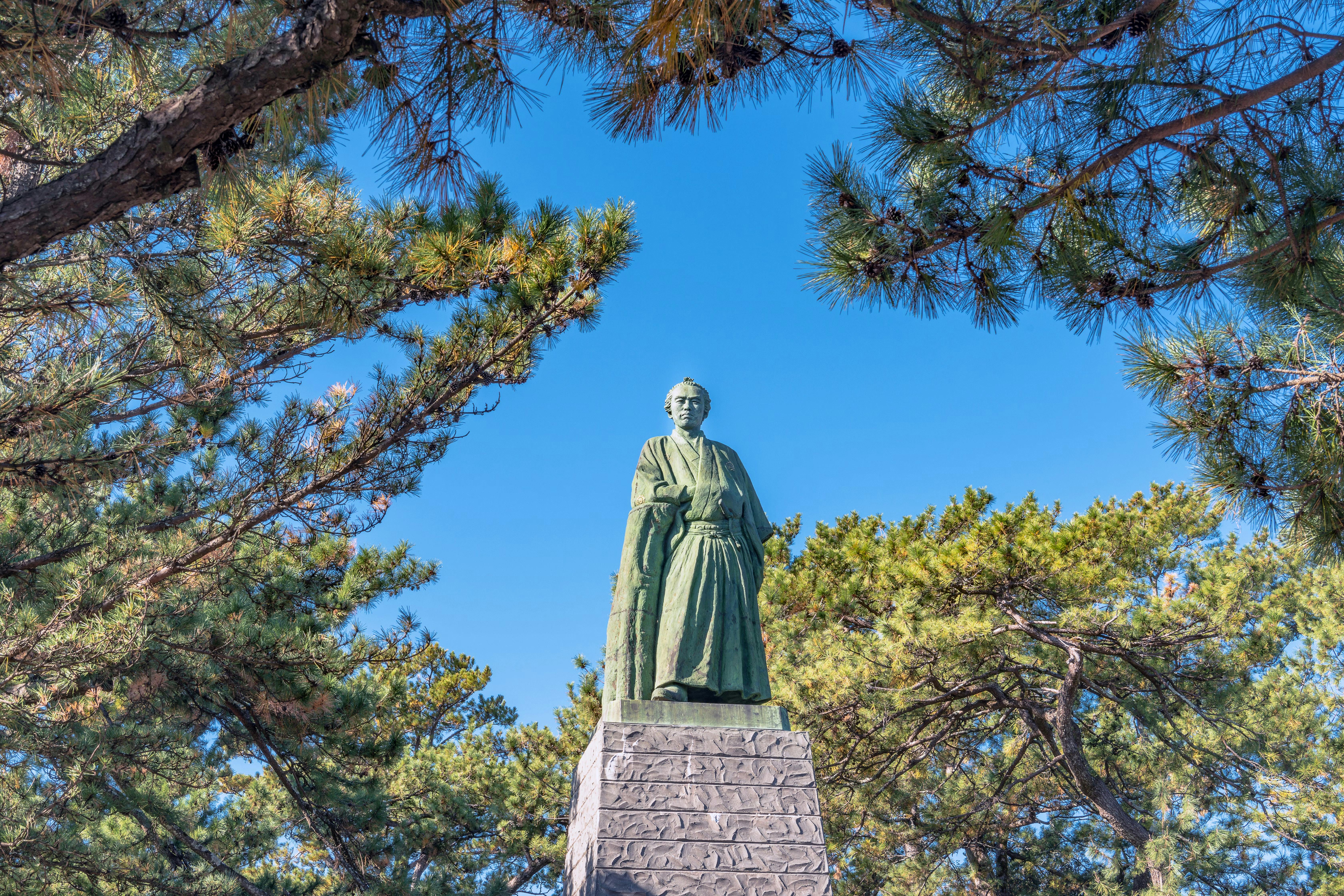 A bronze statue of a person in traditional clothing stands on a stone pedestal, surrounded by green pine trees and set against a clear blue sky.