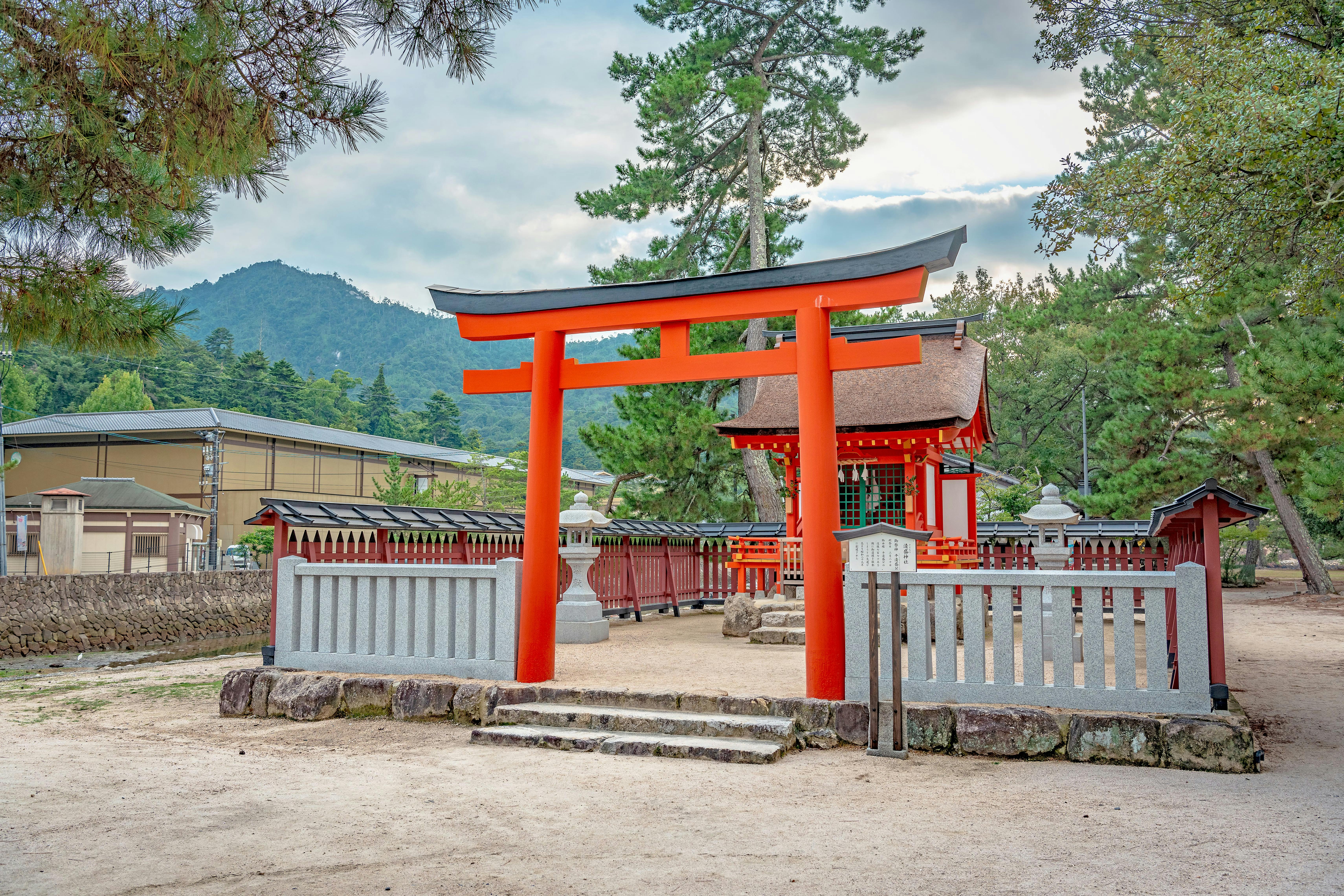 A bright red torii gate stands at the entrance to a traditional Japanese shrine, surrounded by a fence, trees, and mountains in the background under a cloudy sky.