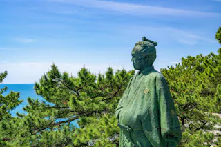 A bronze statue of a man in traditional Japanese attire stands among green pine trees, with a clear blue sky and the sea in the background.