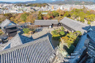 A high-angle view of a traditional Japanese castle courtyard with grey tiled rooftops, manicured gardens, and city buildings, mountains, and colorful autumn trees in the background.