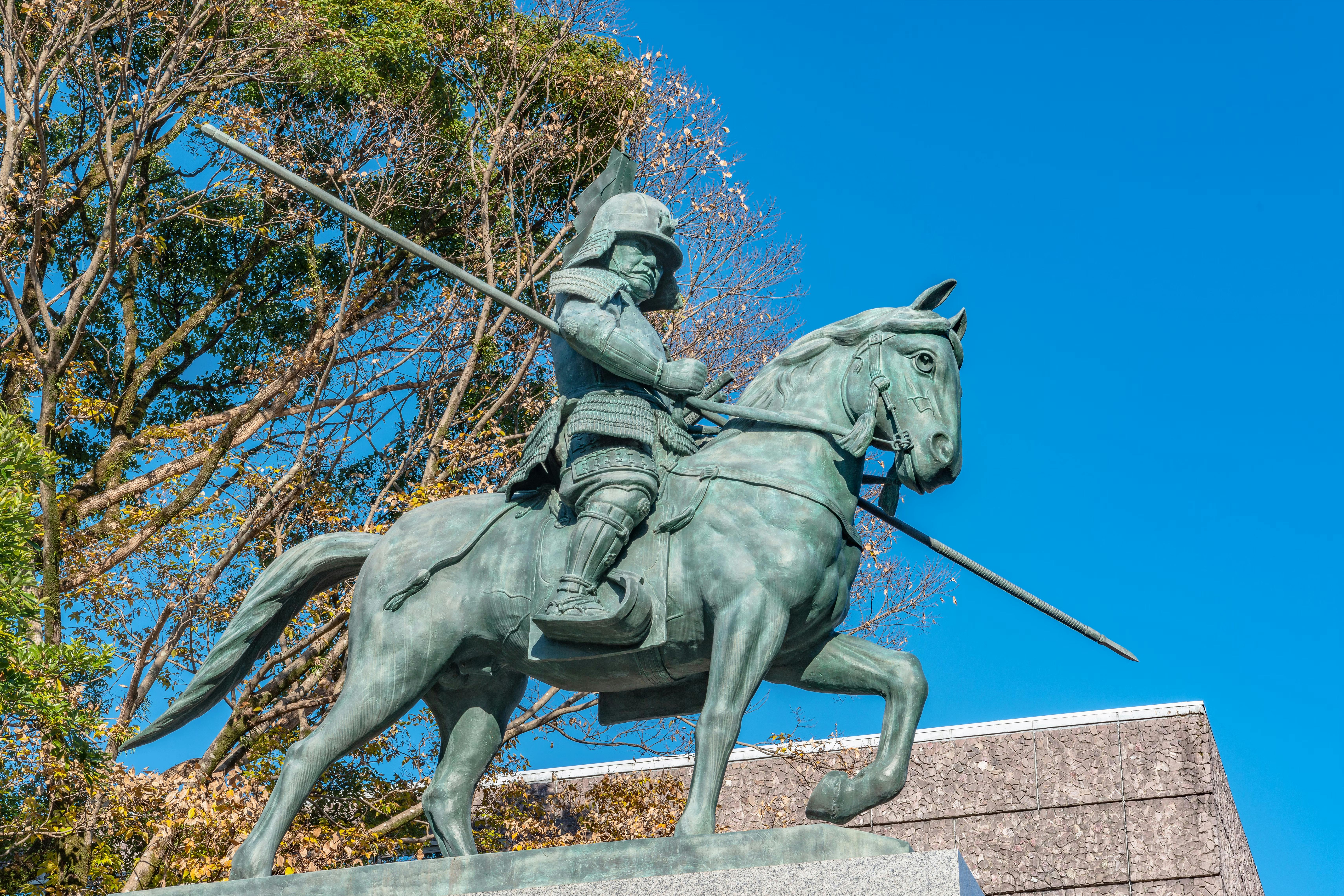 Statue of a samurai warrior in armor riding a horse and holding a spear, set against a clear blue sky with trees and a building in the background.
