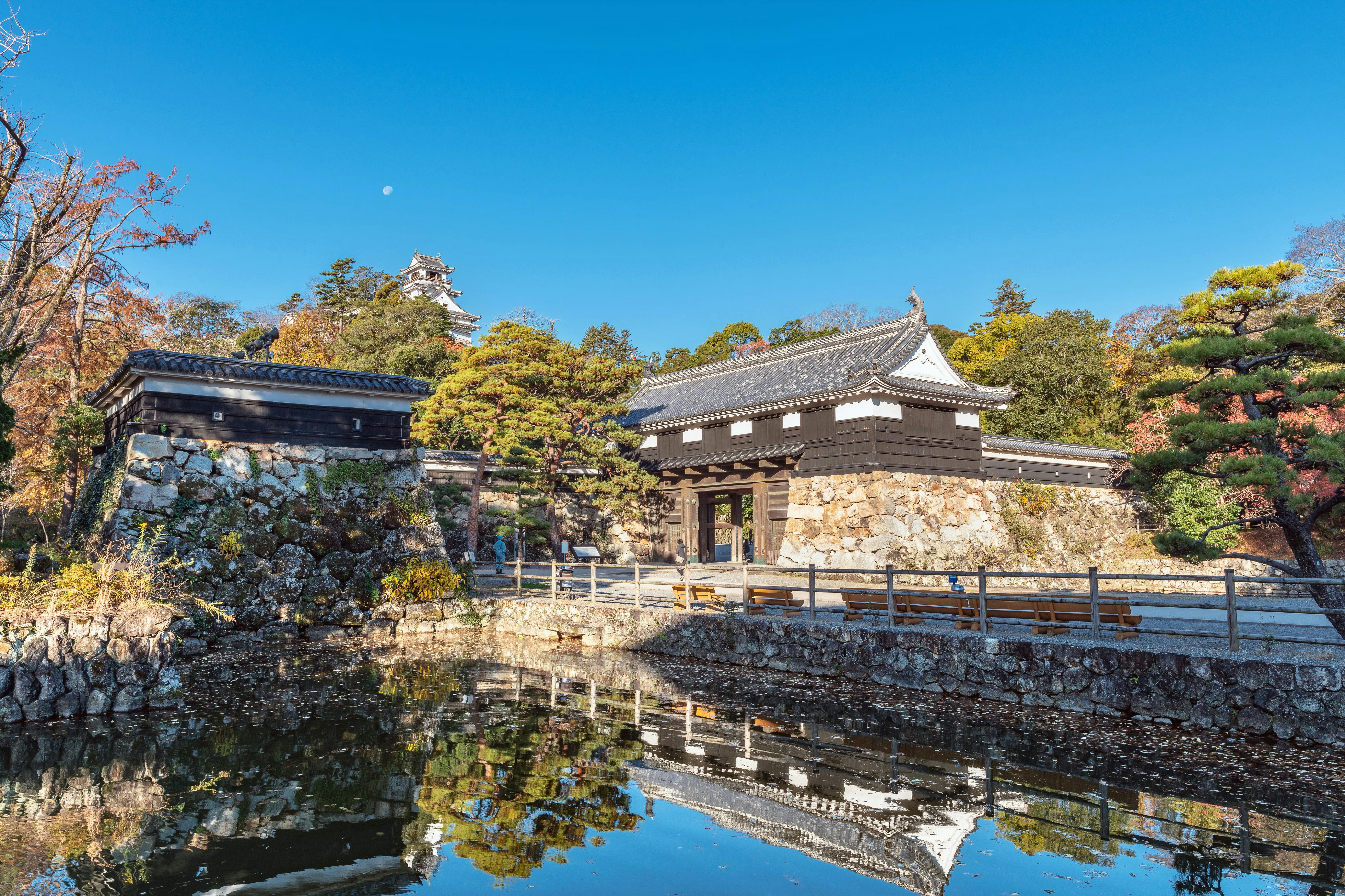 Traditional Japanese castle gate and stone walls reflected in a calm pond, surrounded by autumn trees and greenery under a clear blue sky. A white castle tower is visible in the background.