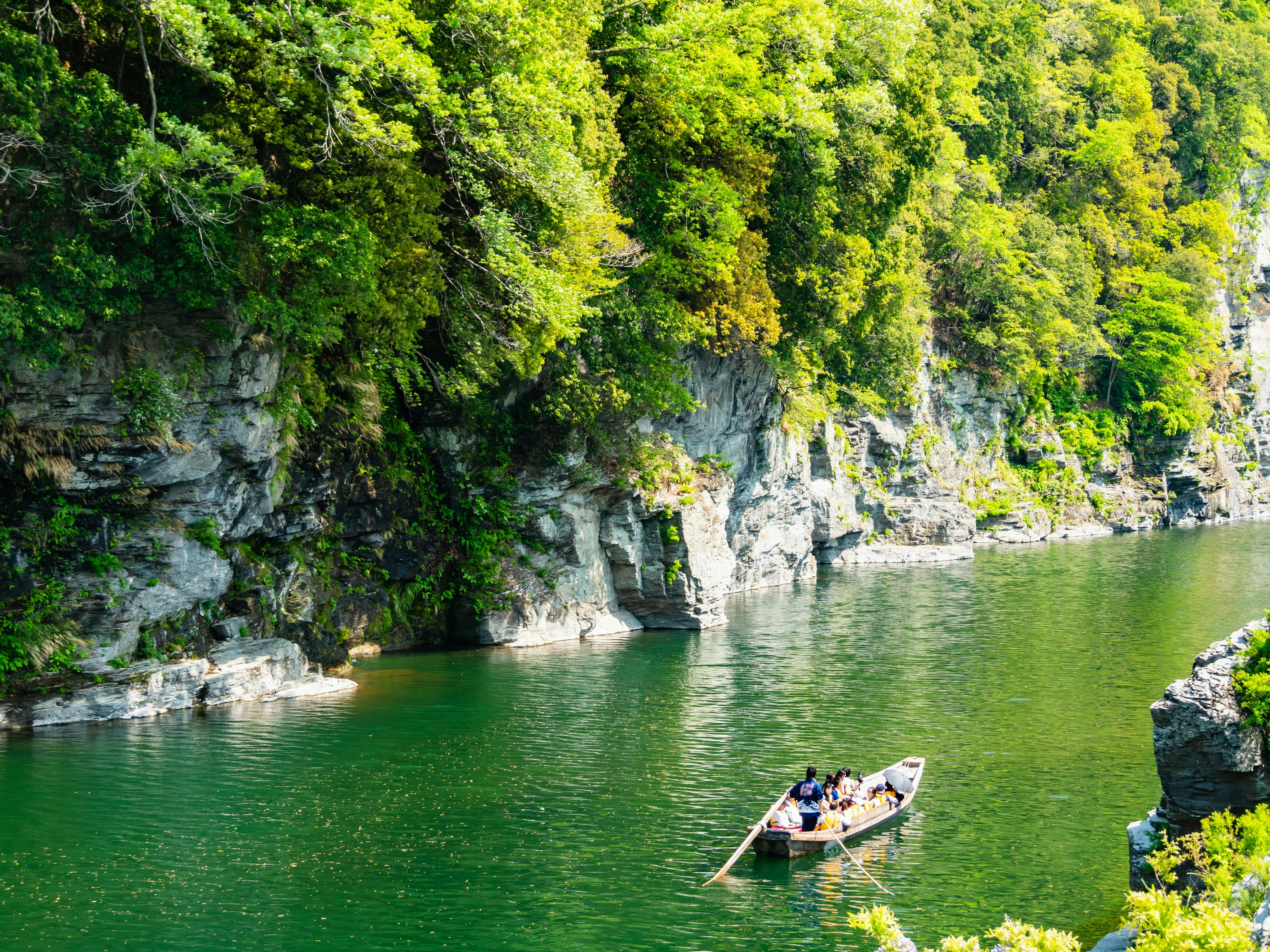 A small wooden boat with several people is rowed by a guide along a green river bordered by steep, rocky cliffs and lush, leafy trees on a sunny day.