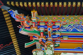 Colorful, ornate wooden architectural detail of a Japanese temple, featuring intricate patterns, geometric designs, and a vibrant dragon carving beneath a black and gold roof.