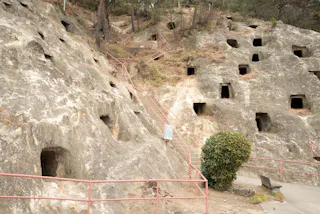 Rocky hillside with multiple square cave openings, metal railings, a staircase ascending the slope, and a bush near a bench at the bottom right.