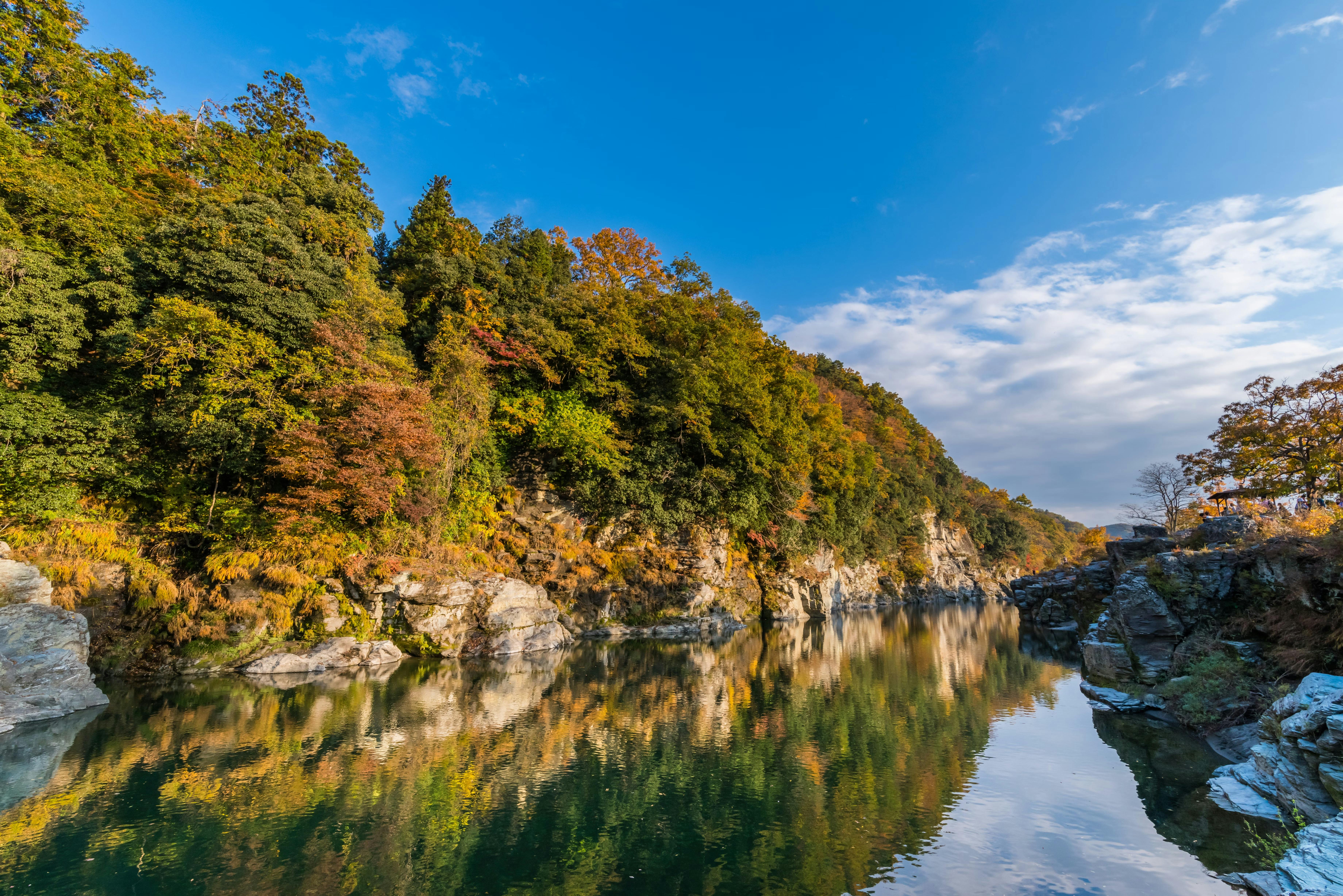 A calm river reflects a forested hillside with autumn foliage under a blue sky with scattered clouds. Rocky banks line the water, and the sunlight highlights the vibrant colors of the trees.
