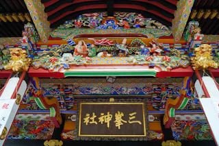 Colorful, ornate wooden carvings and detailed decorations adorn the entrance of a traditional Japanese shrine, with a wooden sign featuring kanji characters hanging at the center.