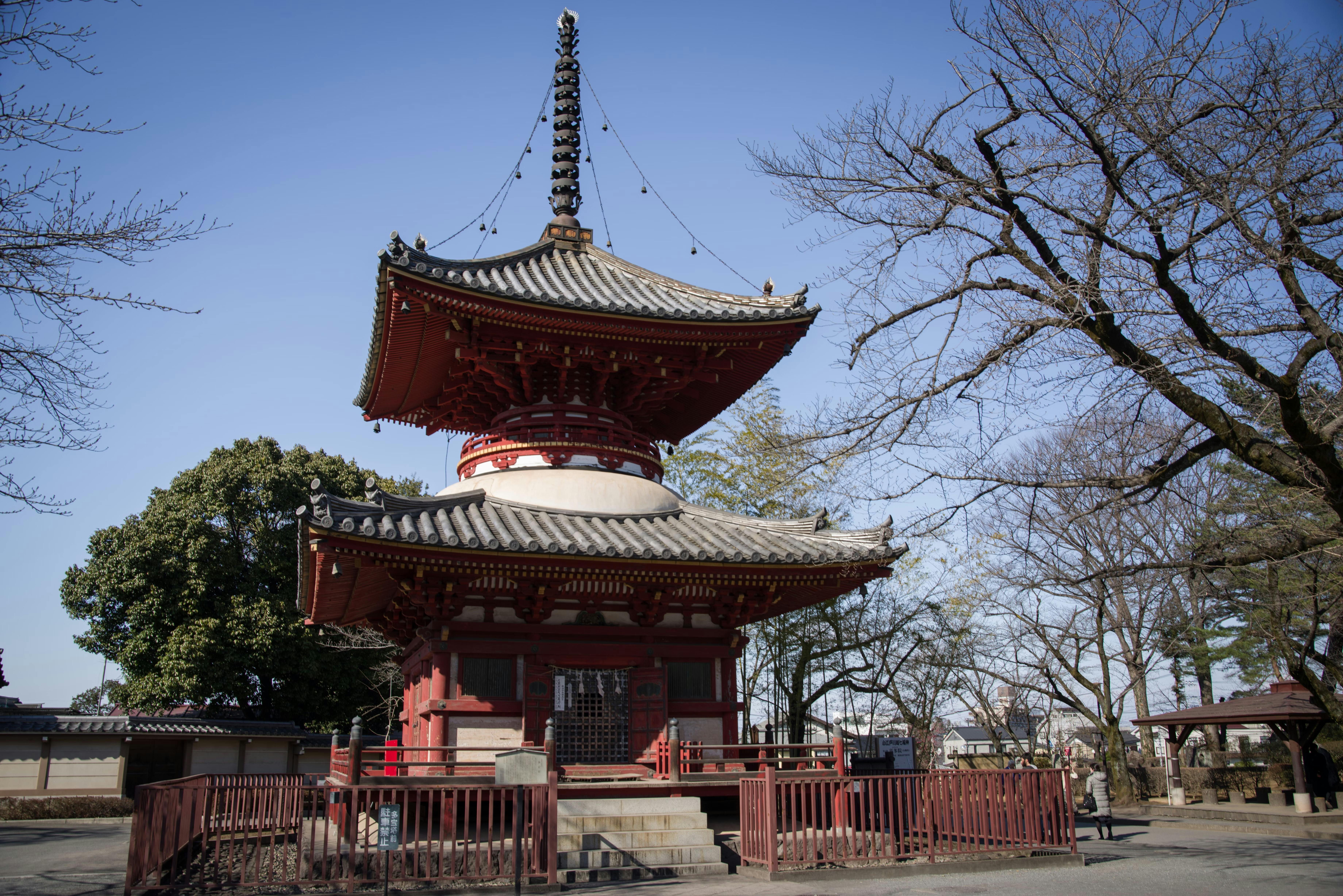 A traditional Japanese pagoda with a tiled roof and intricate wooden details stands surrounded by bare trees and a fence on a clear, sunny day.