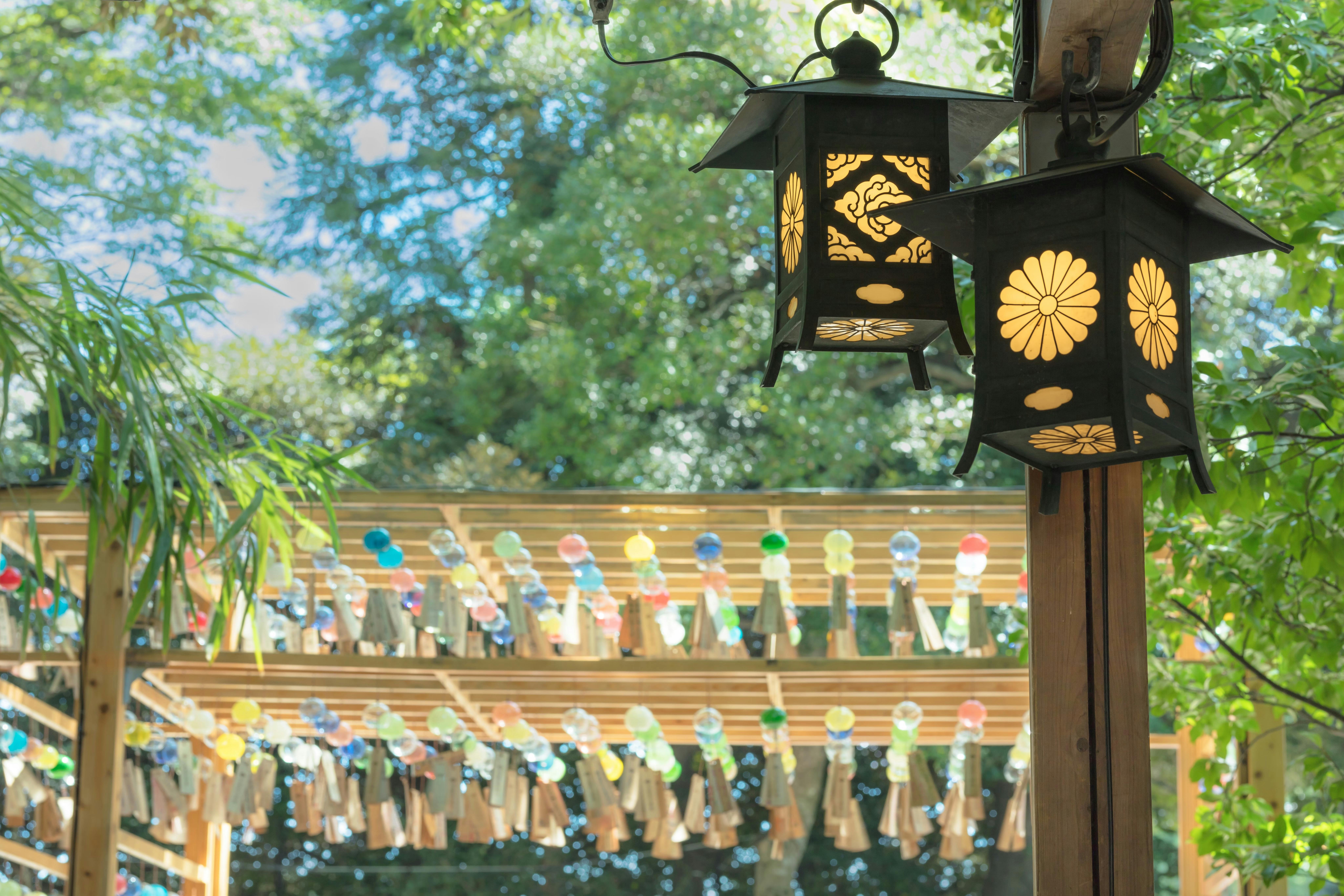 Two black decorative lanterns hang in the foreground, each with a floral pattern. In the background, colorful glass wind chimes are displayed on wooden frames amid lush green trees.