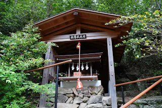 A small wooden Shinto shrine with a slanted roof stands among green trees, with a rope and bell hanging in front, and stone steps leading up to the entrance.