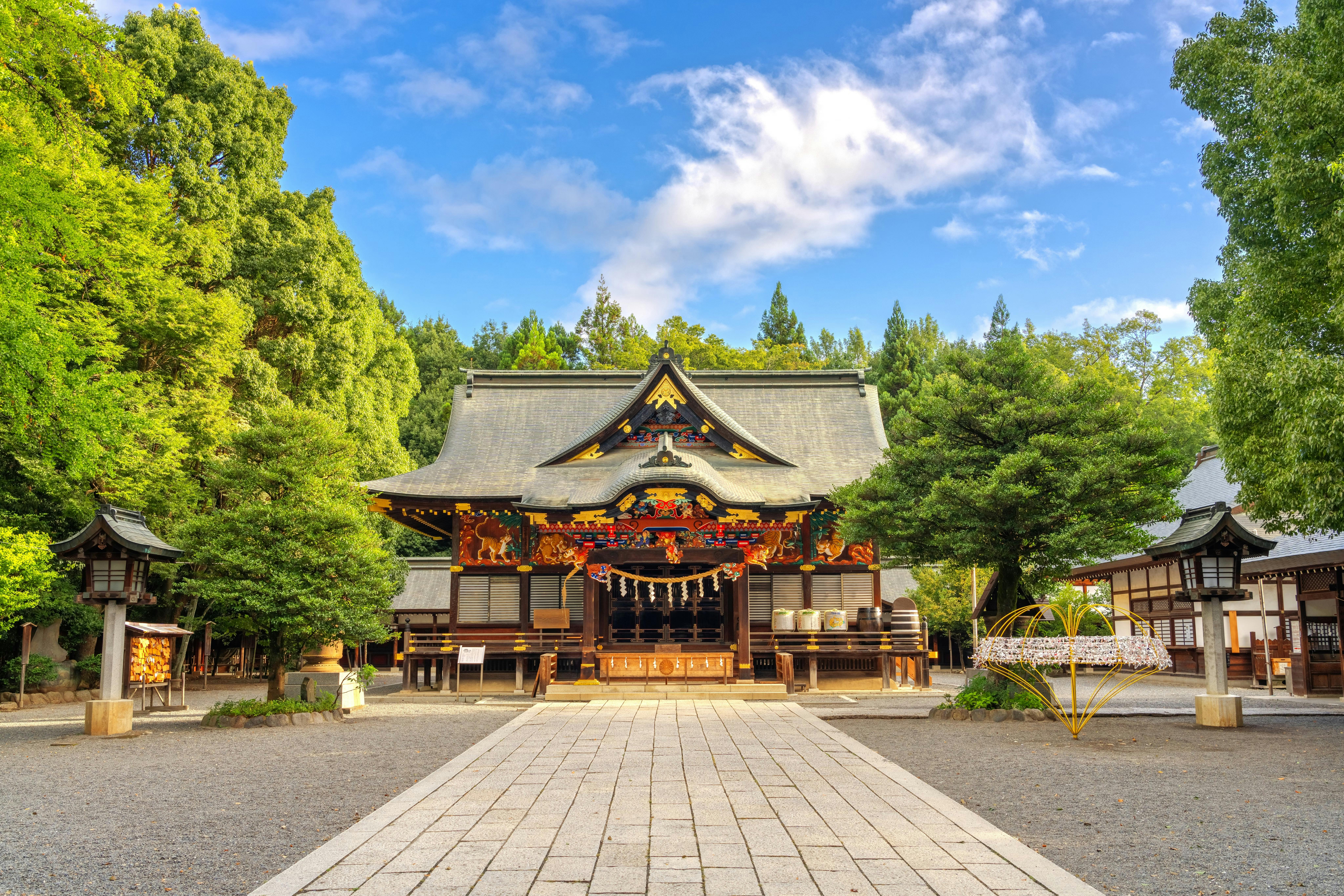 A traditional Japanese Shinto shrine with ornate wooden architecture, surrounded by lush green trees under a bright blue sky with scattered clouds. A stone path leads to the shrine’s entrance.