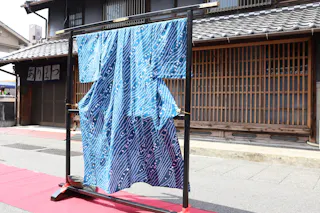 A blue kimono with a white wave and dot pattern hangs on a black display stand outdoors, in front of a traditional Japanese wooden building with tiled roof and sliding lattice doors.