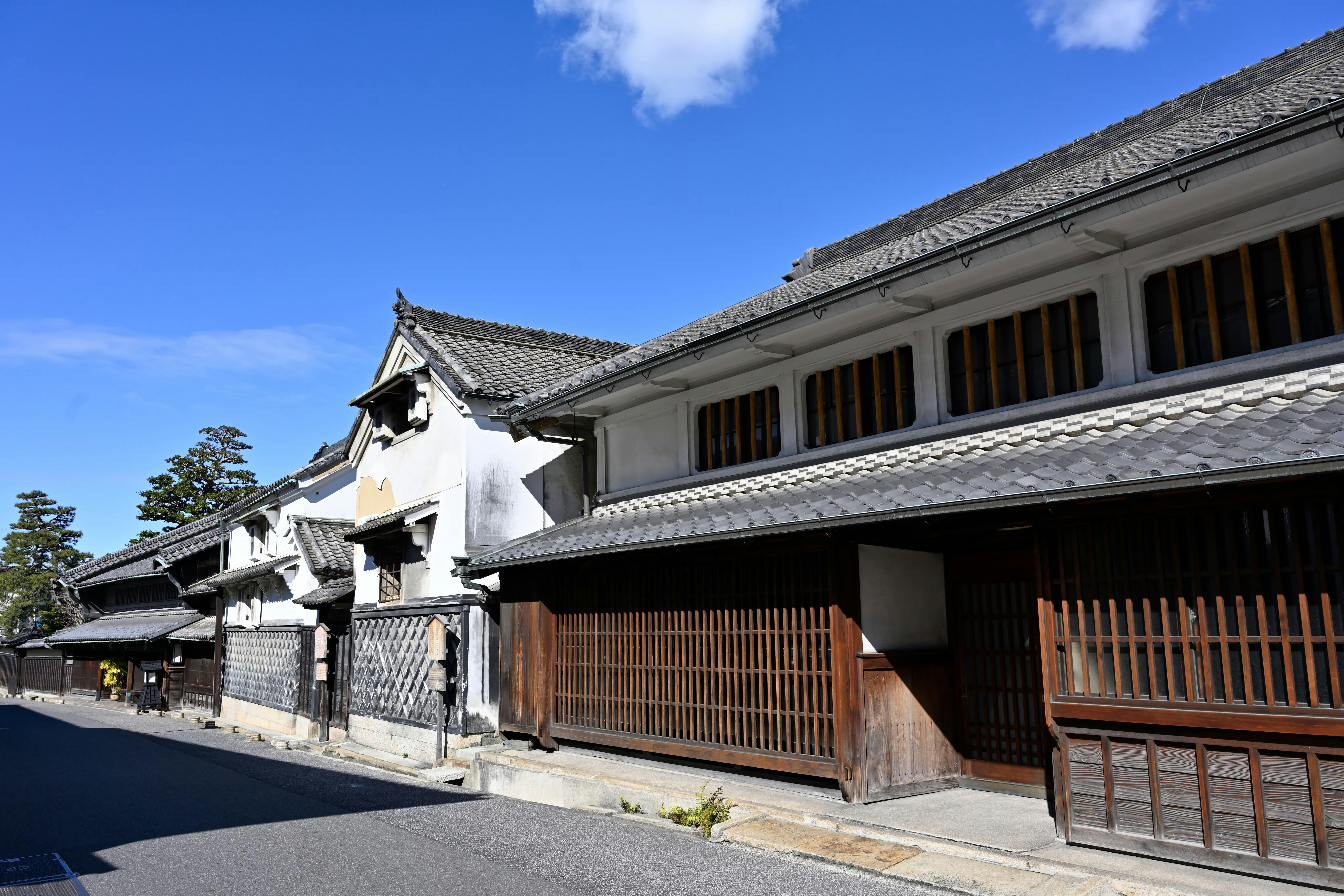 Traditional Japanese wooden buildings with tiled roofs line a quiet street under a clear blue sky, showcasing classic architectural features such as wooden lattice doors and white plaster walls.