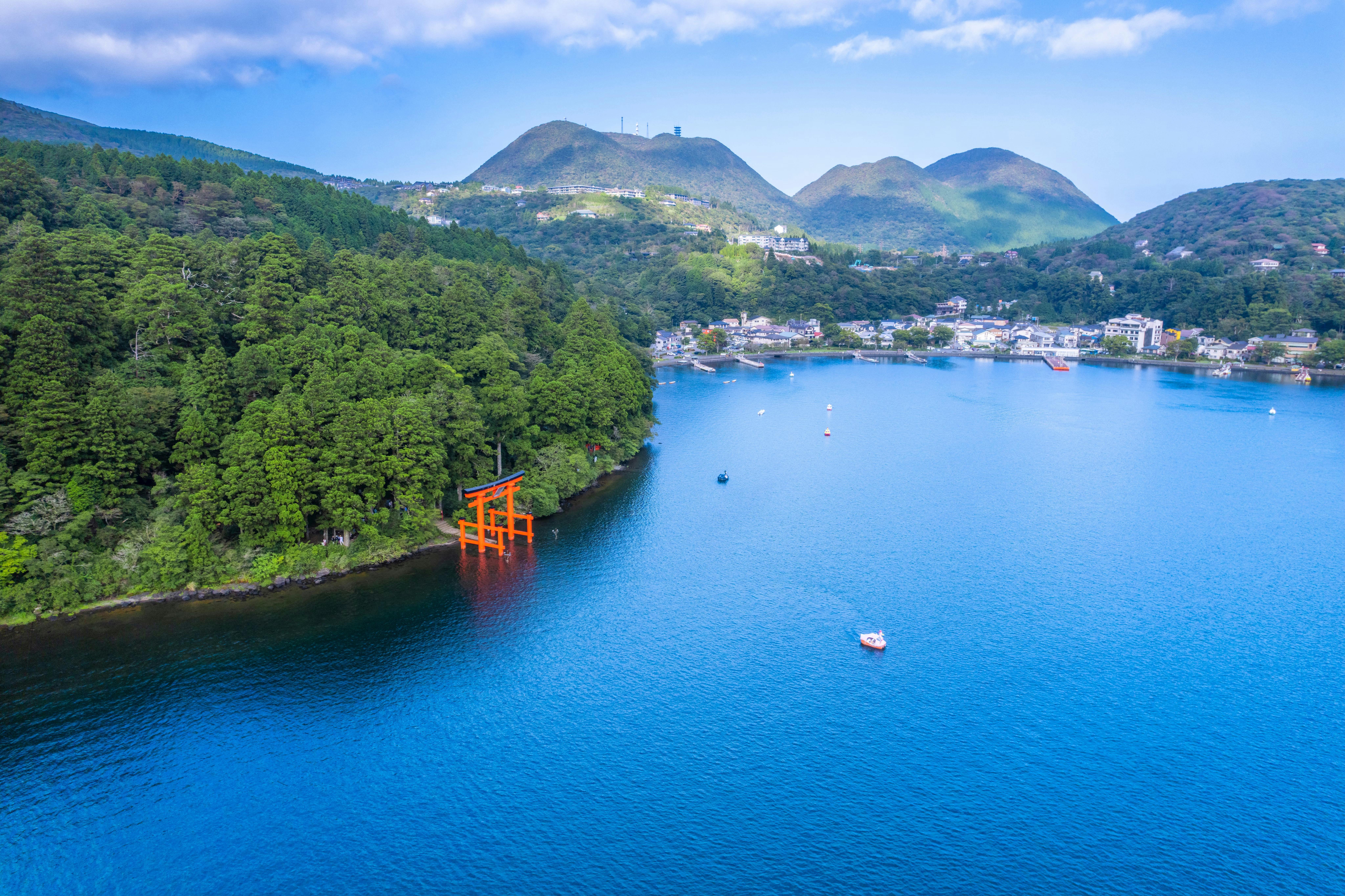 Aerial view of a large blue lake with a red torii gate near a forested shoreline, distant mountains, and scattered buildings; a few boats are on the water under a partly cloudy sky.