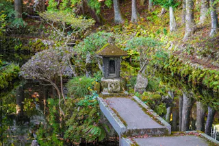 A stone lantern stands on a moss-covered pedestal at the end of a narrow path over a reflective pond, surrounded by lush greenery and tall trees in a tranquil garden setting.