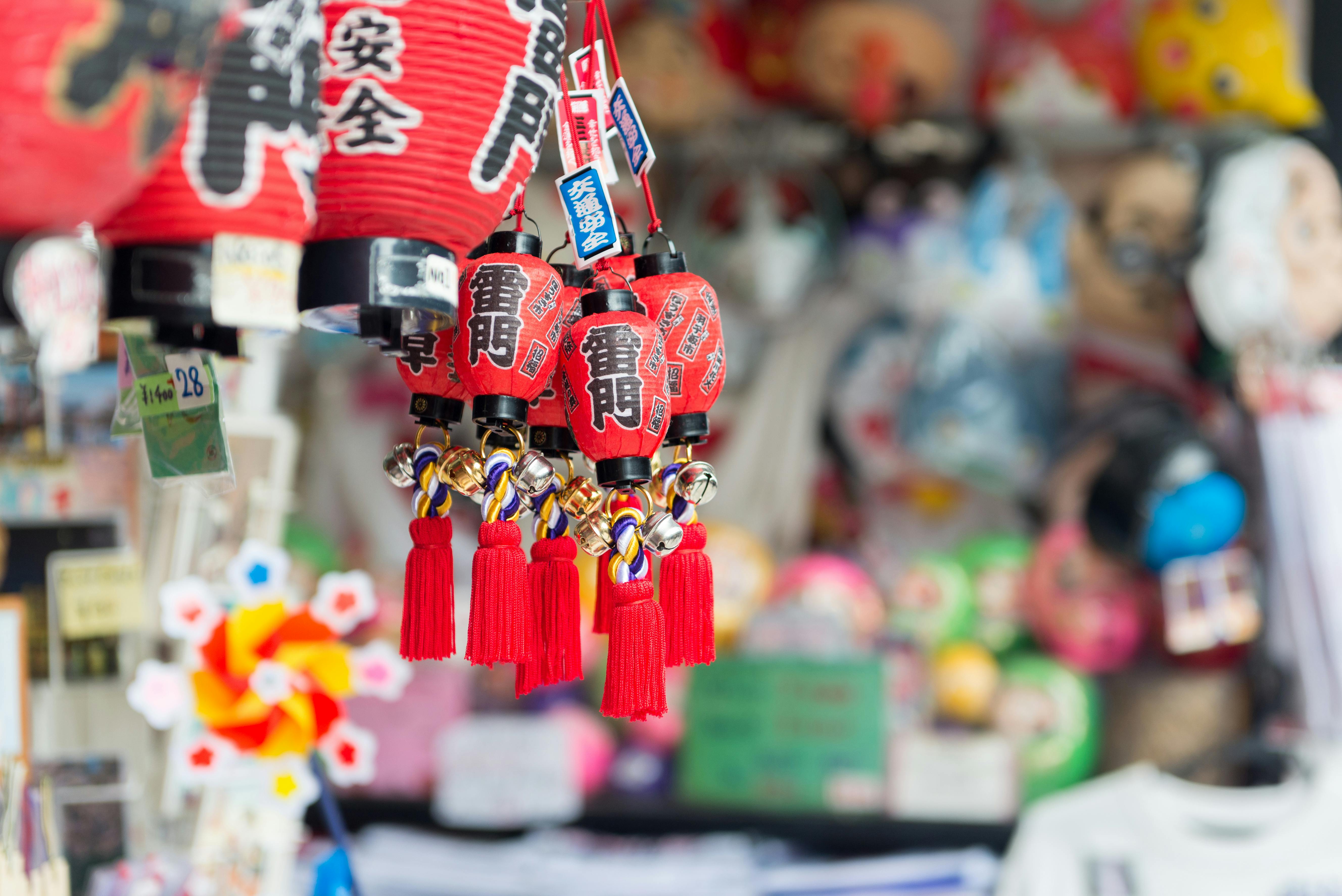 Red Japanese lantern keychains with gold and purple cords and tassels hang in focus at a street market stall, with colorful blurred souvenirs and plush toys in the background.