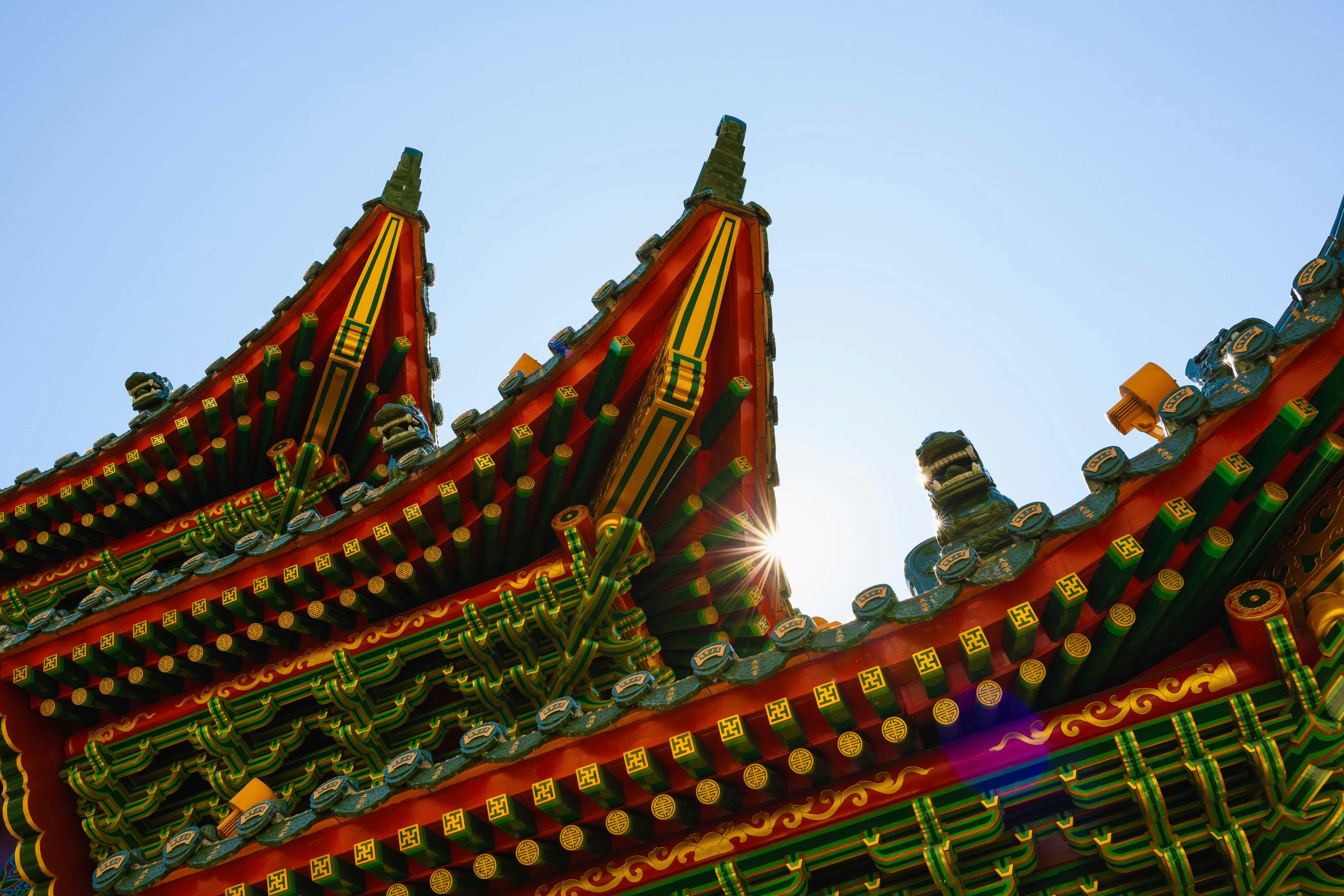 Colorful, ornate rooftops of a traditional Asian temple with intricate patterns and vibrant green, yellow, and red details, set against a clear blue sky with sunlight peeking through the eaves.
