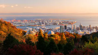 Cityscape of Kobe, Japan at sunset, with colorful autumn trees in the foreground, urban buildings, and the harbor with docks and cranes beside the calm sea. Mountains and a gradient sky are in the background.
