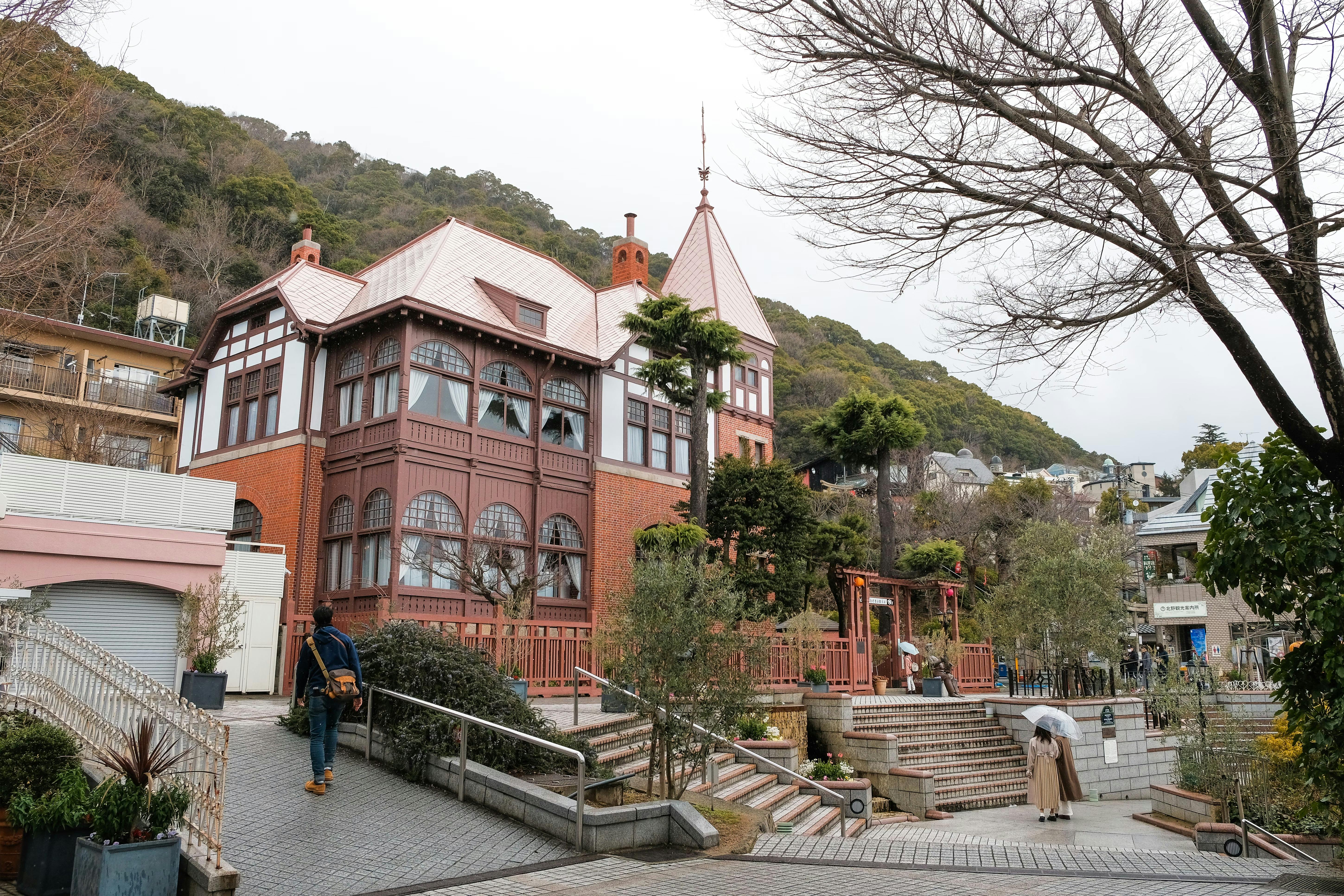 A red-brick, European-style house with large windows and a tall spire sits on a sloped street. Trees surround the building, and a few people walk nearby. Hills covered in greenery rise in the background.