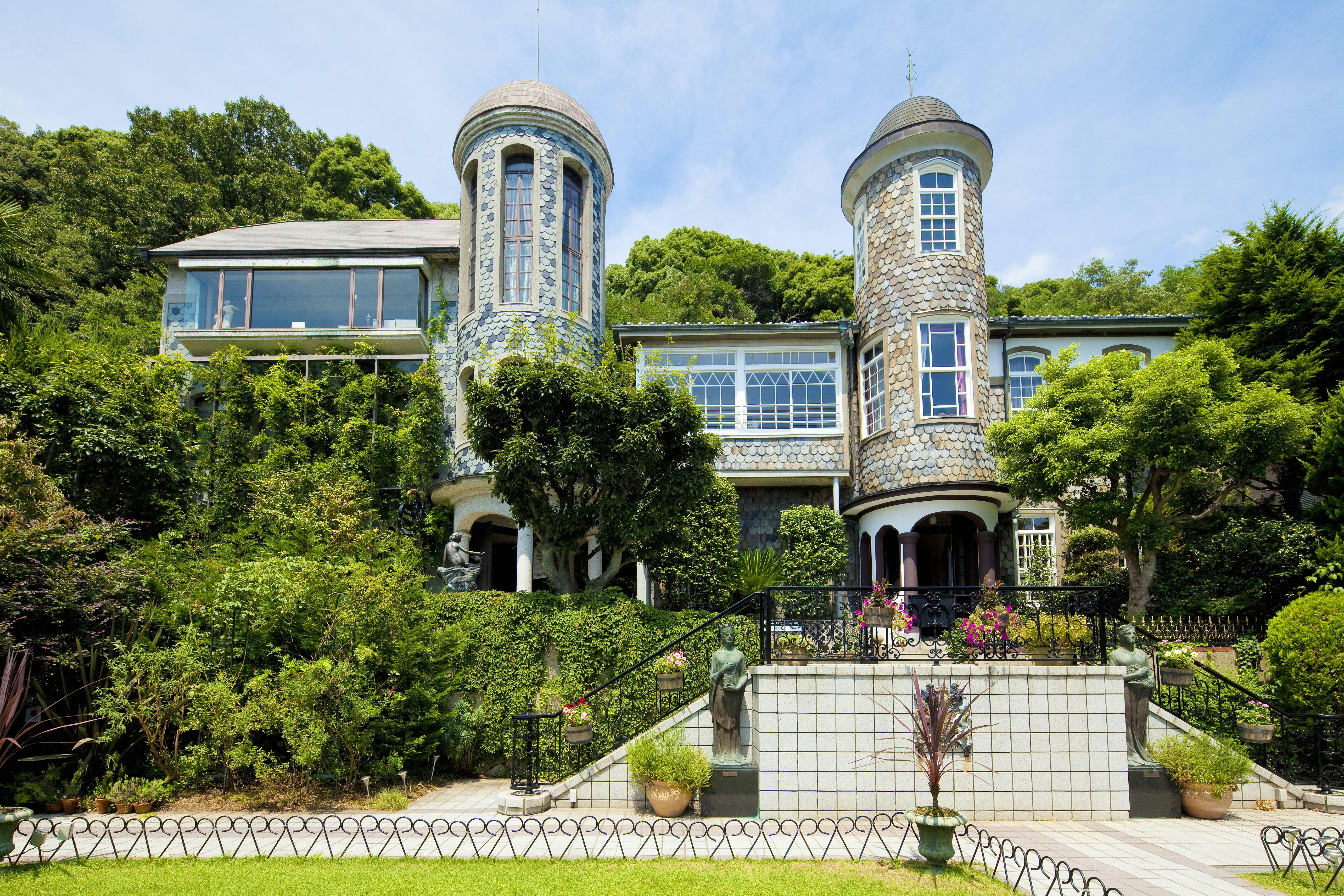 A large, elegant house with two round towers, arched windows, and ivy climbing the stone walls, surrounded by lush greenery and a landscaped garden under a blue sky.