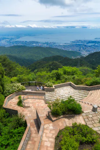 A scenic overlook with two people sitting on a bench, surrounded by lush green hills, offers a panoramic view of a coastal city and the sea under a partly cloudy sky.