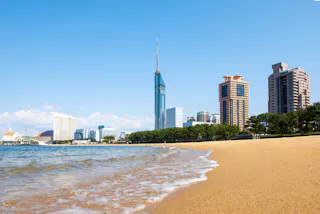 A sandy beach with gentle waves in the foreground, modern high-rise buildings and a tall tower in the background under a clear blue sky.