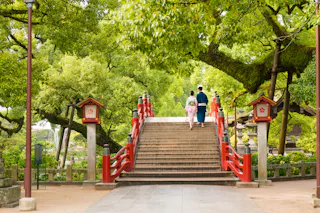 Two people in traditional Japanese clothing walk up a red bridge surrounded by lush green trees, with lanterns on either side of the bridge.