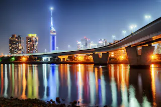 A brightly lit cityscape at night with a tall illuminated tower, modern buildings, and a bridge spanning over calm water reflecting colorful city lights.