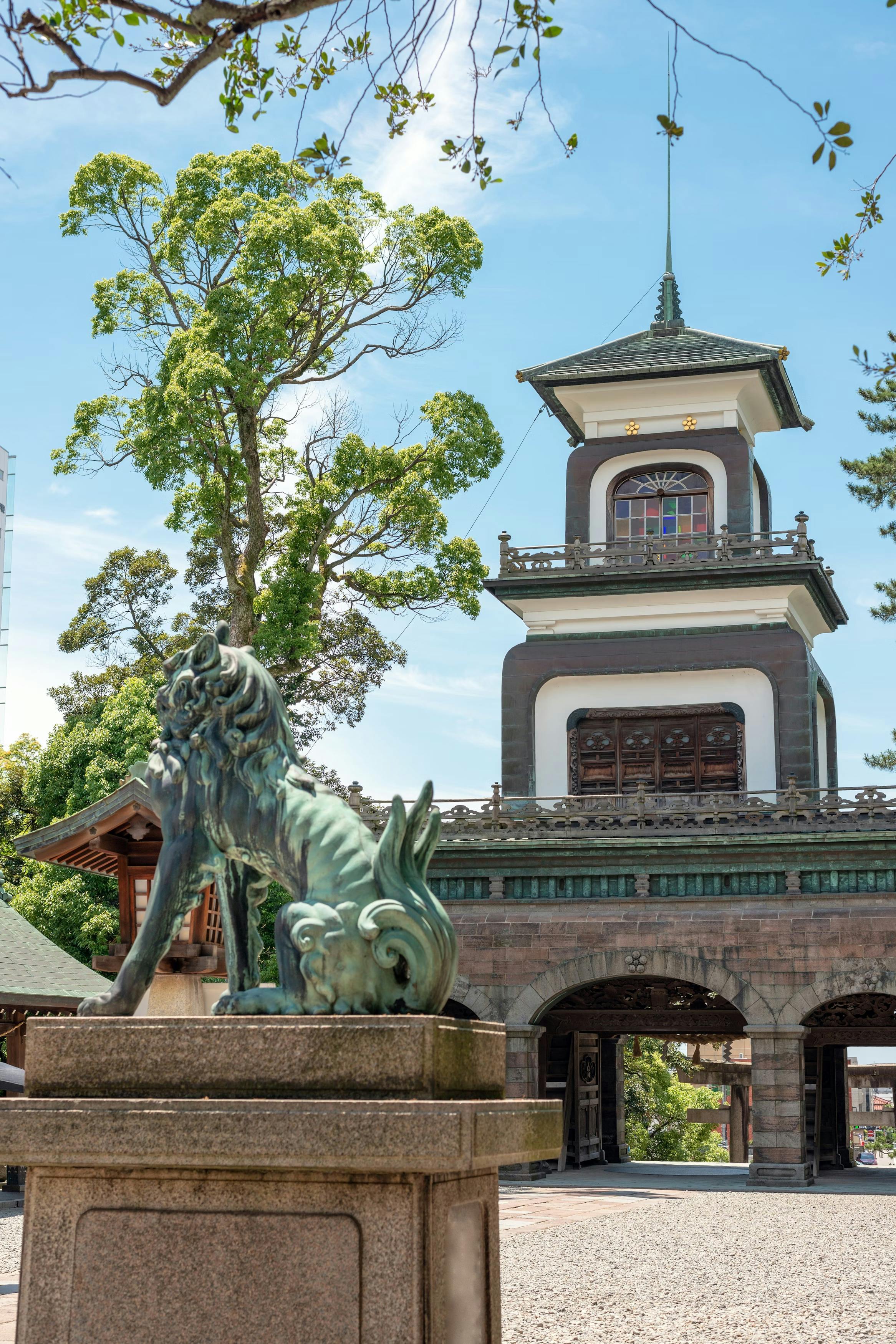 A bronze lion-dog statue stands on a stone pedestal in front of a historic Japanese gate with a tower, surrounded by trees under a bright blue sky.