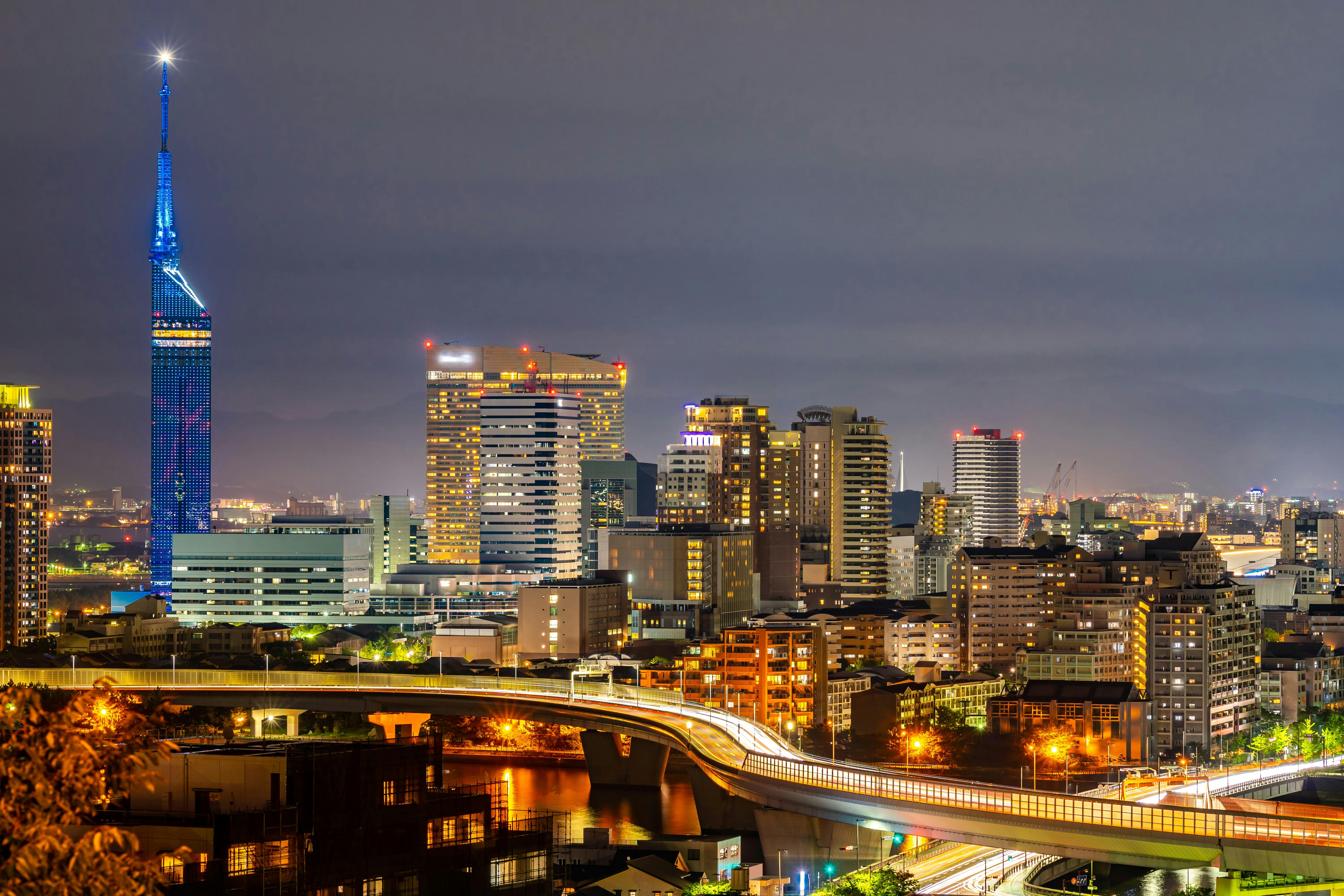 A cityscape at night featuring a brightly lit tower with blue lights on the left, surrounded by illuminated modern buildings and highways with streaks of car lights in the foreground.